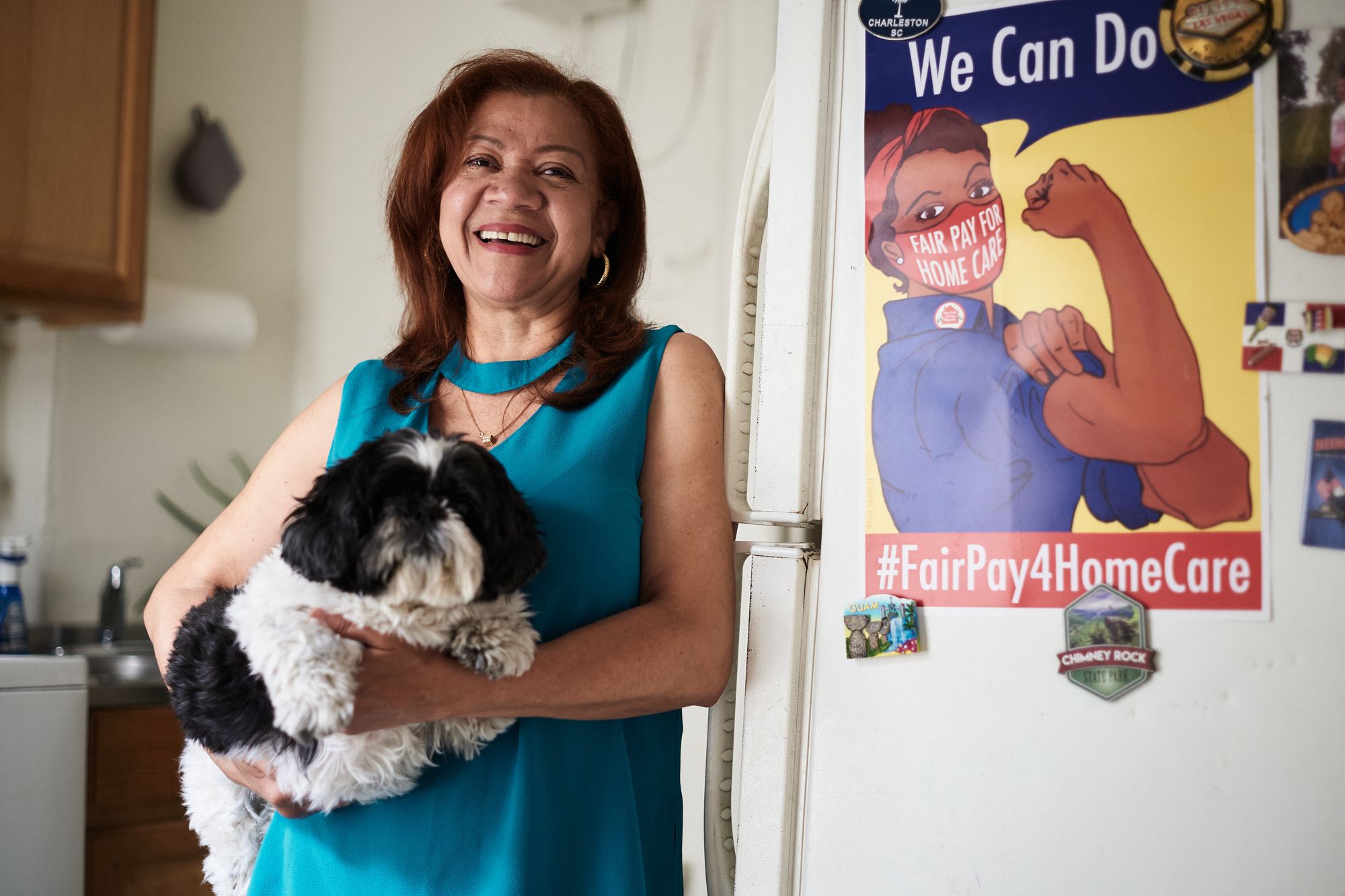 Calvo standing in her apartment in front of a fridge with a "#FairPay4HomeCare" sign while holding her dog.