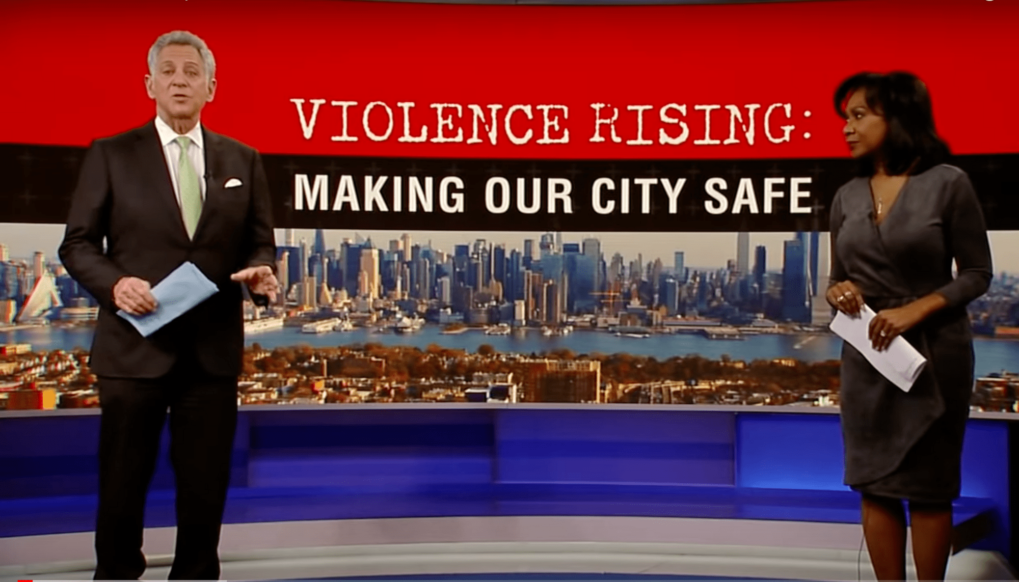 Local news anchors stand in front of a greenscreen emage that reads: "Violence Rising: Making Our City Safe"