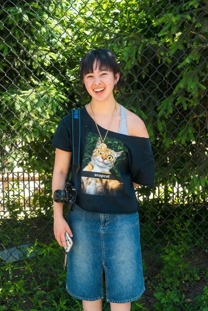 Bassett wears a black T-shirt with a wide-eyed cat and the lettering "snapcat" with long blue jean shorts. She's holding her phone with a camera slung over her right shoulder.