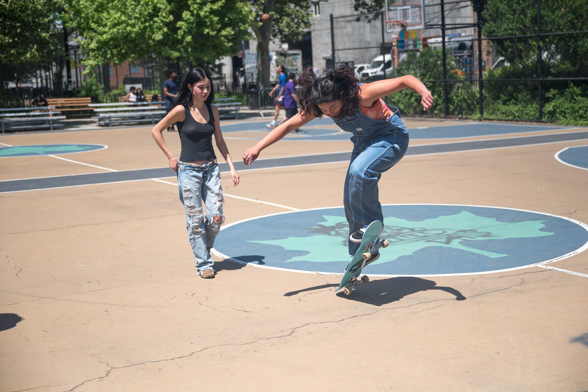 One woman doing a trick on a skateboard while another one watches at a park in Brooklyn.