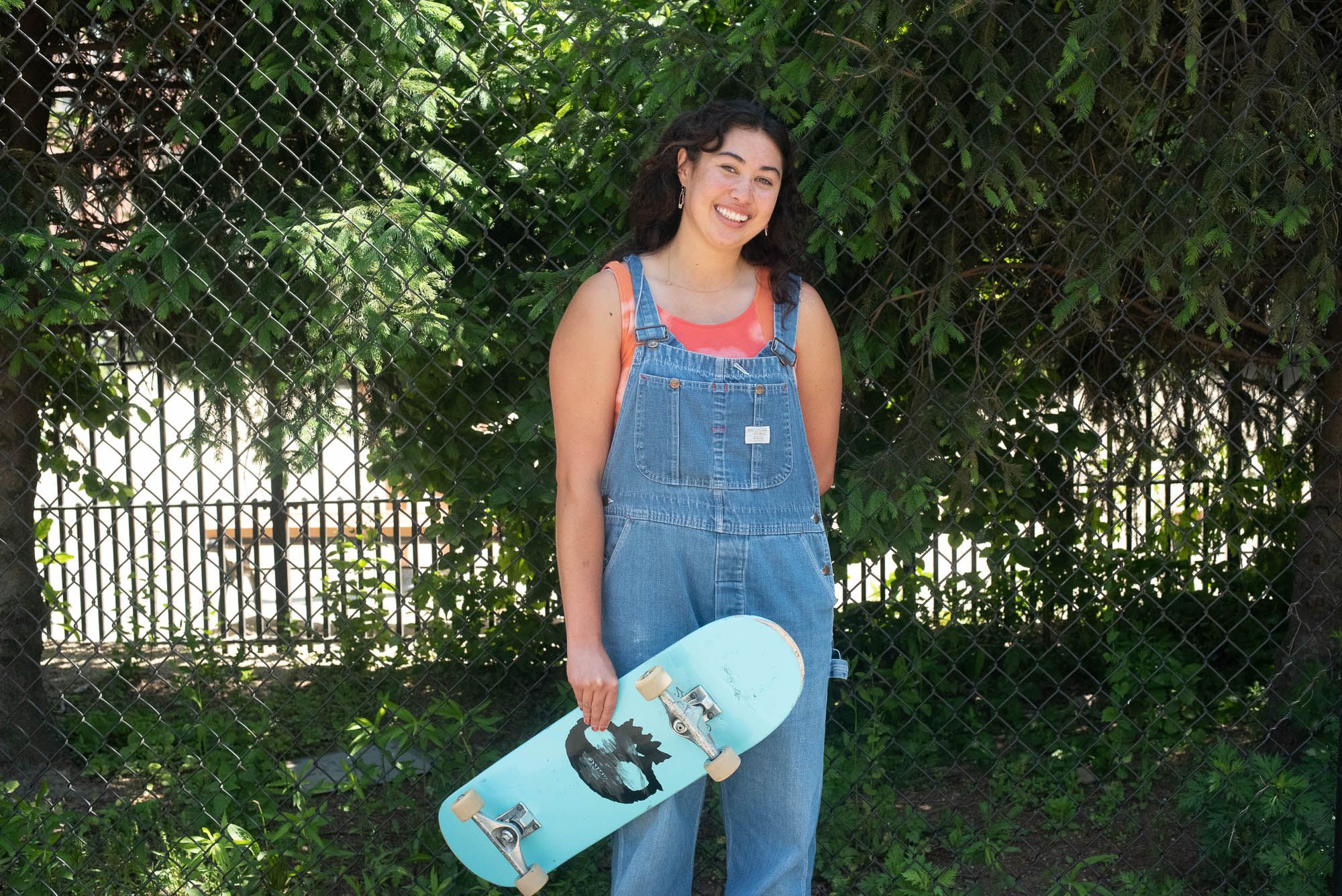 Mullin smiles at the camera wearing blue jean overalls and an orange top underneath while holding her light blue skateboard with a black skull on it.