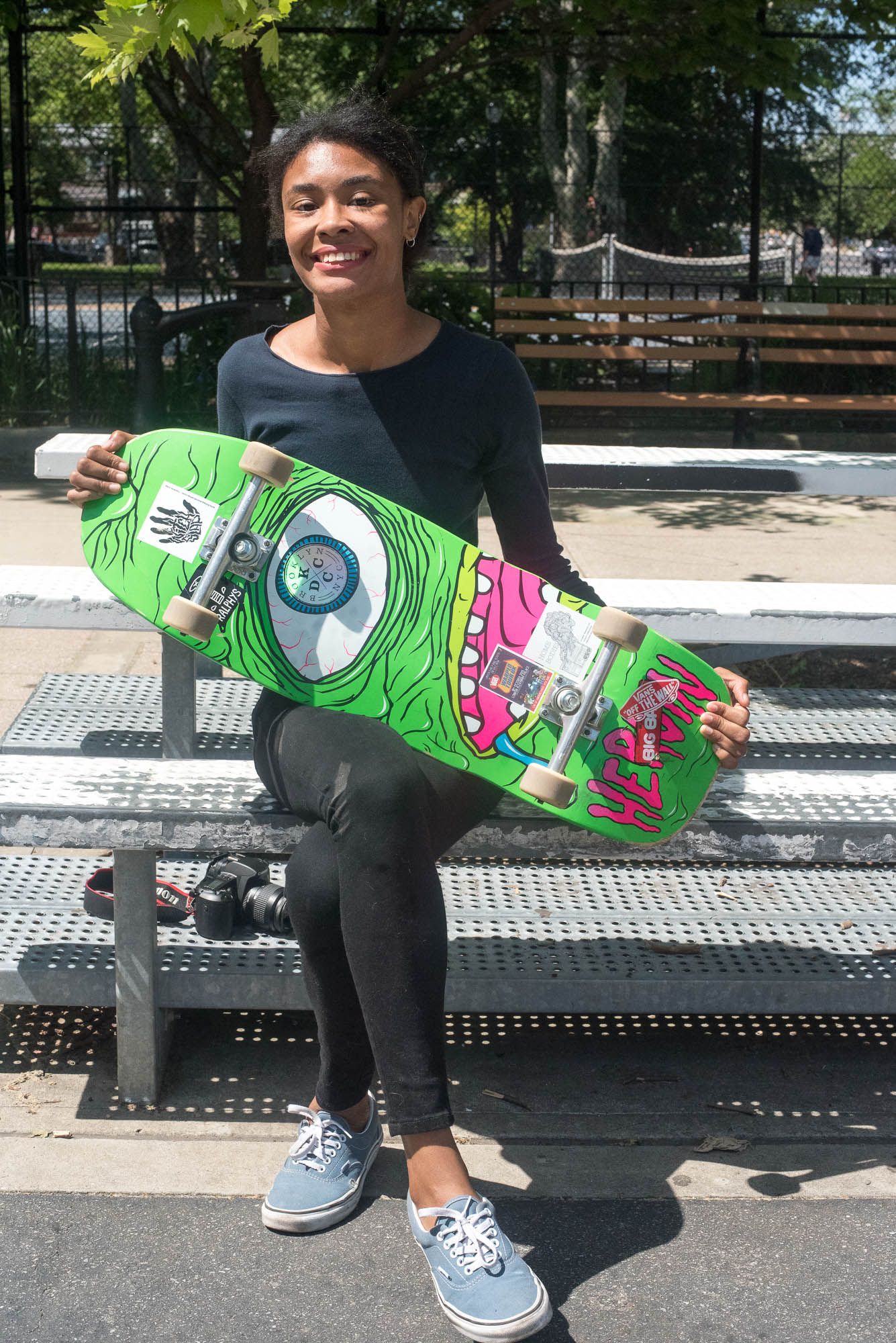 Moore wears all black, sitting on the bleachers showing off her skateboard that has a lime green and pink alien-like creature on it..