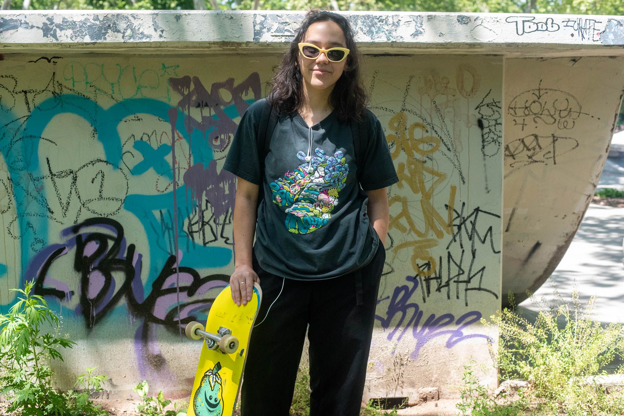 Paxman with her left hand in her pocket wearing black clothing and yellow sunglasses next to her skateboard, which is bright yellow with a blue-green fruit that's smiling.