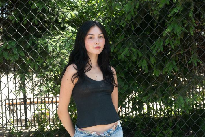 Bryan stands facing the camera with her arms tucked behind her back wearing a black tank top and blue jeans.