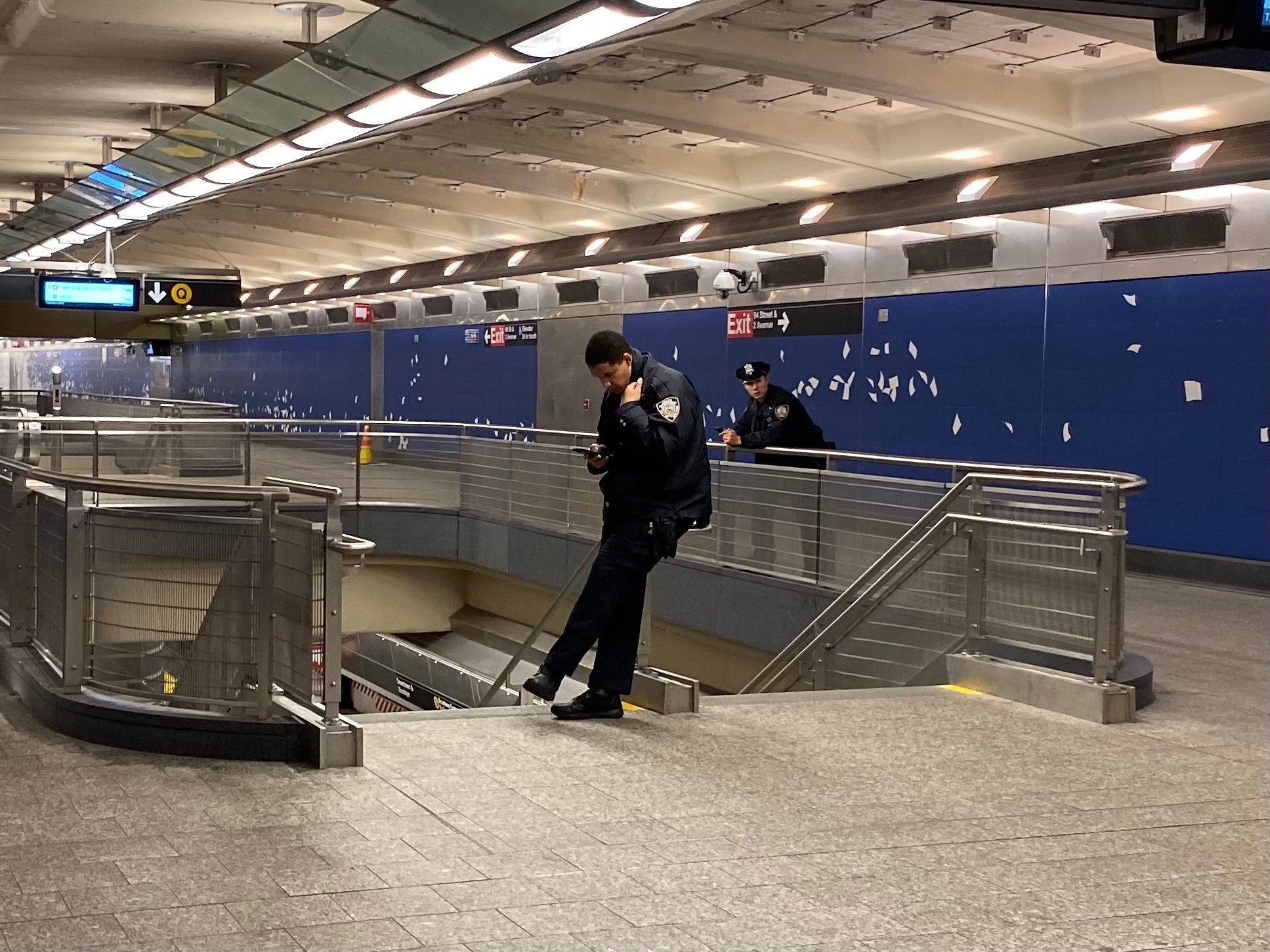 An NYPD officer atop a subway station stairwell looks at his phone while another NYPD officer behind him looks on