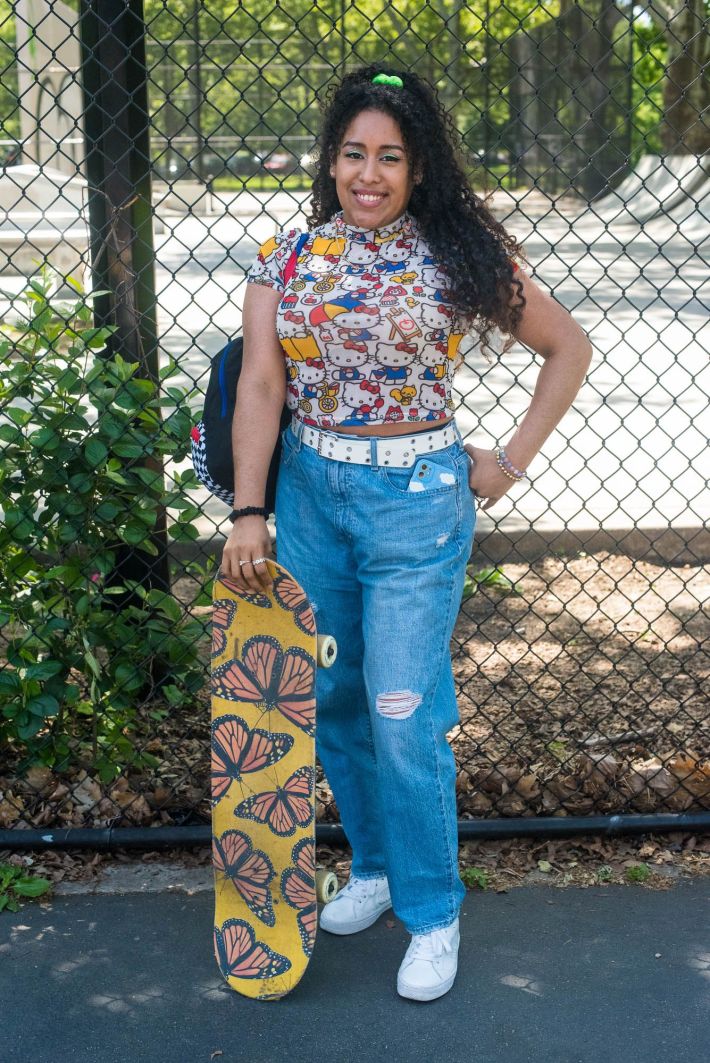 Trochez standing with a Hello Kitty top, blue jeans, white sneakers, curly hair and a lime green scrunchie. Her skateboard has a Monarch butterfly design.