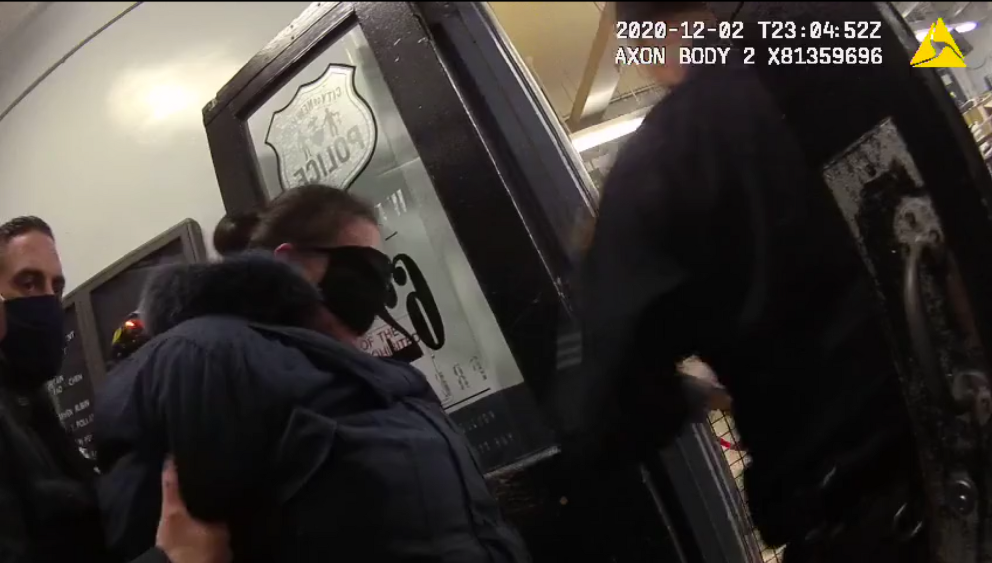 A handcuffed woman in a winter coat is led through a police precinct door with her mask pulled up over her eyes.