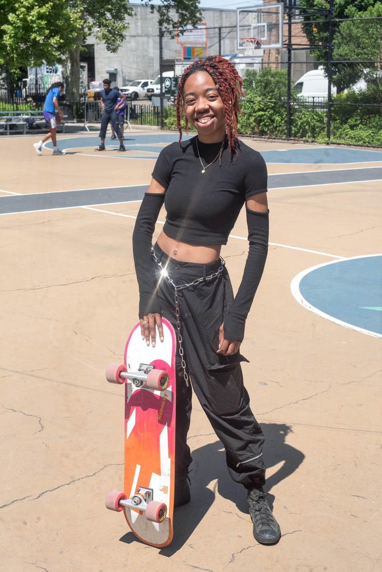 Vision-Skye with red braids, wearing all black next to a pink and orange skateboard. Her chain glistens in the sun as she smiles at the camera.