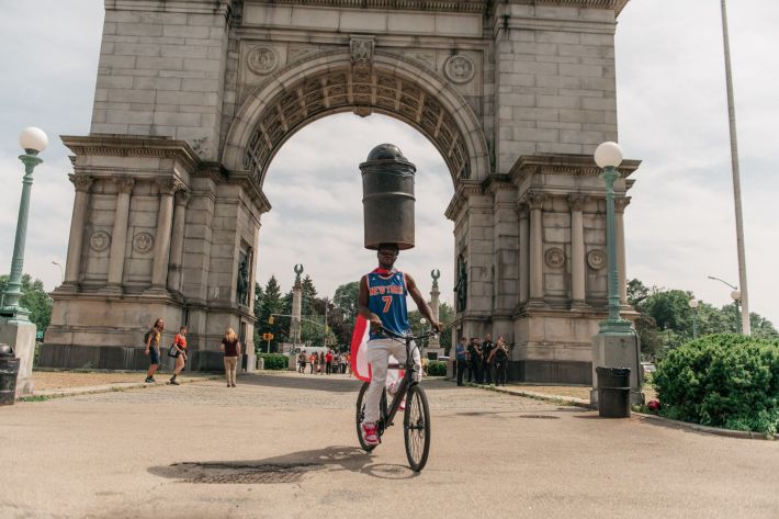 Leh-Boy balances a trash can on his head while riding a bike under the Grand Army Plaza arch