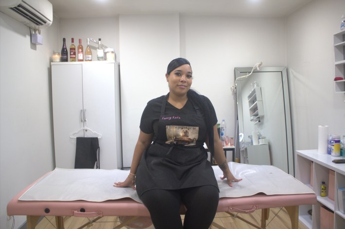 Tisha Gloss, in a black T-shirt, pants, and apron, sits on a pink table in a room of her waxing salon in the Bronx.