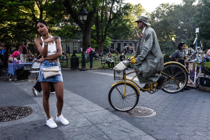 A performer in a plaid suit rides a yellow bicycle near a woman wearing a denim mini skirt.