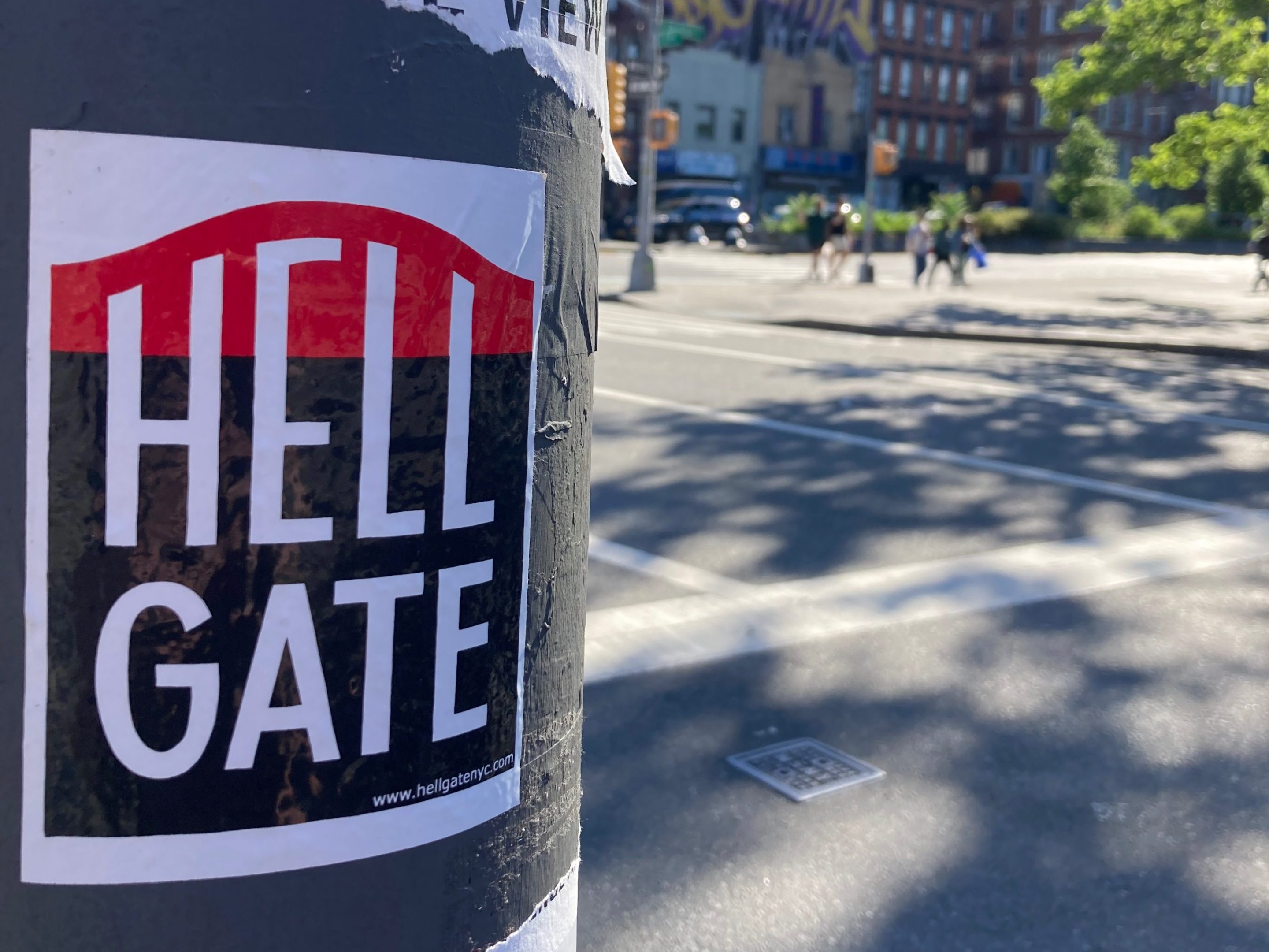 A Hell Gate Sticker on a lamppost in front of a sun-dappled city playground