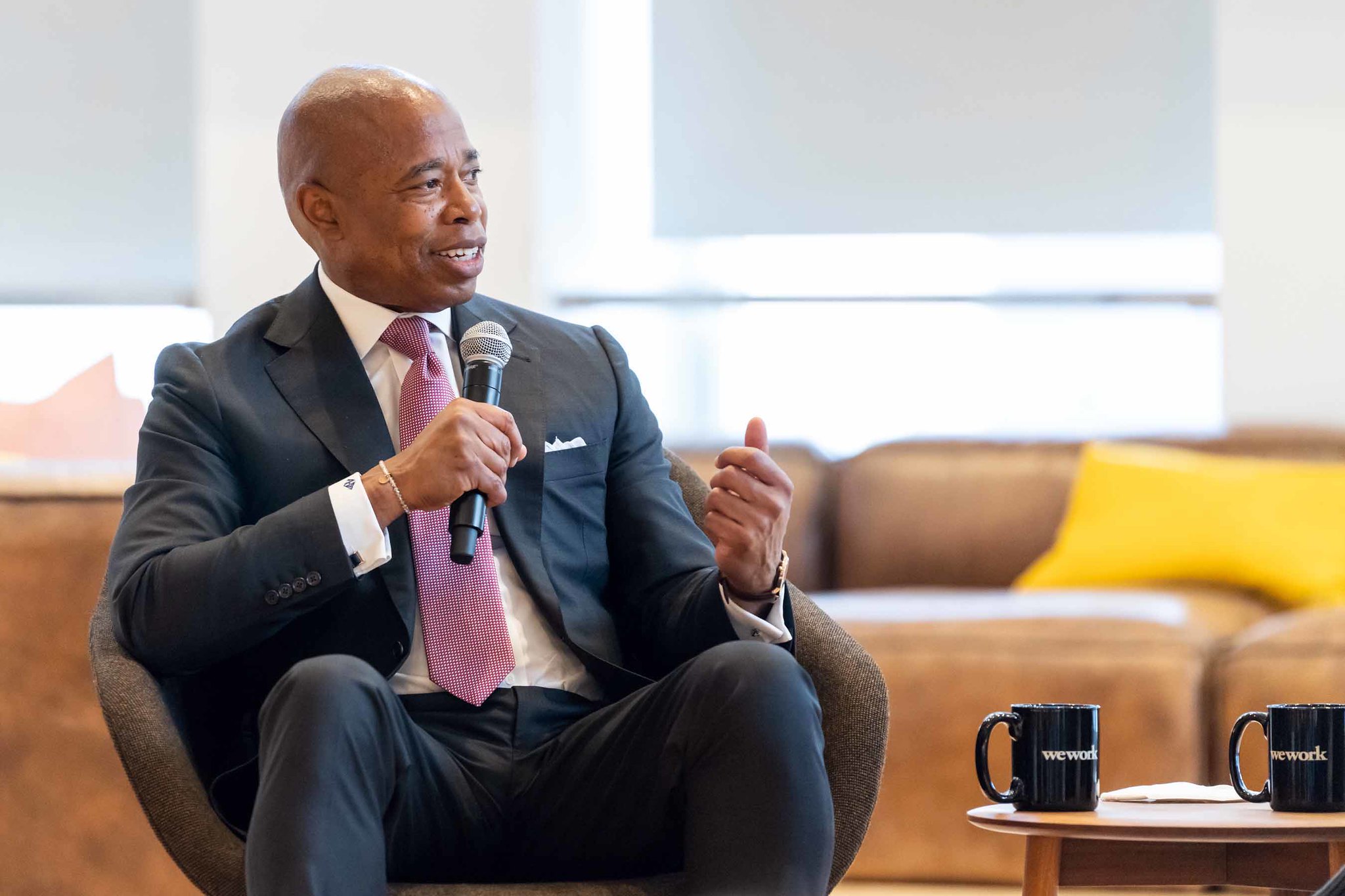 Mayor Eric Adams wears a grey suit and a pinkish tie and holds a microphone while he talks at a conference held at a WeWork.