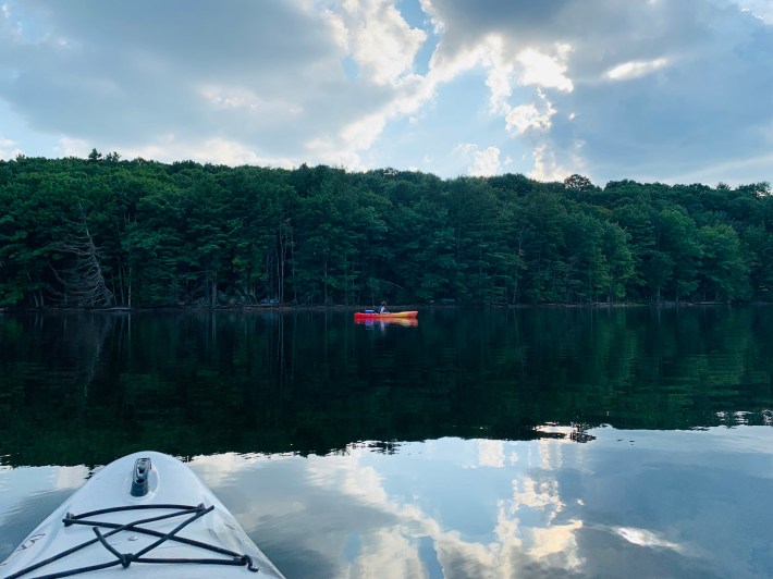 A red, yellow, and orange kayak floats on the Neversink Reservoir, with a backdrop of hemlock trees.