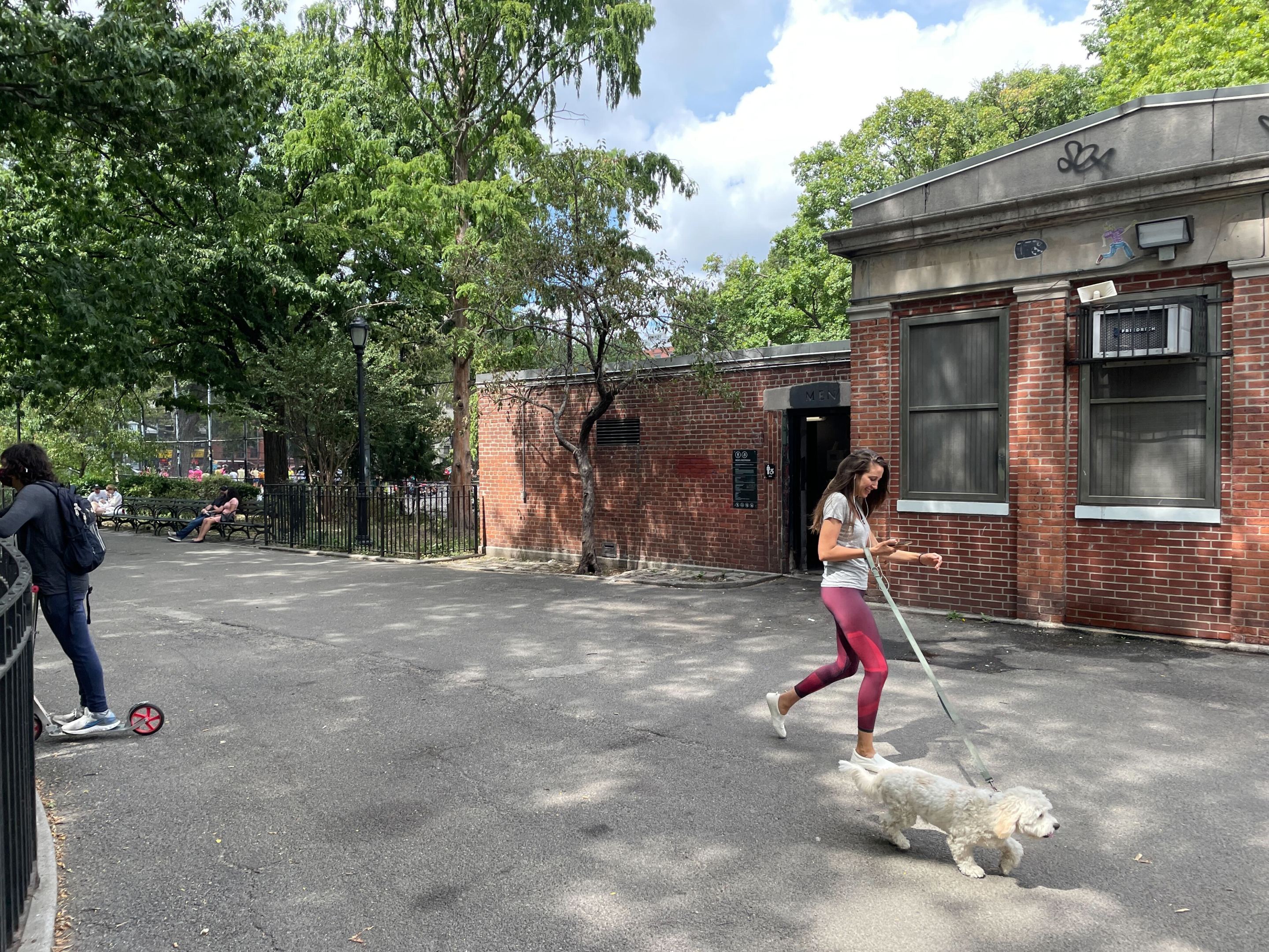 A woman walks a white fluffy dog in front of the Tompkins Square Park bathrooms on a summer day.