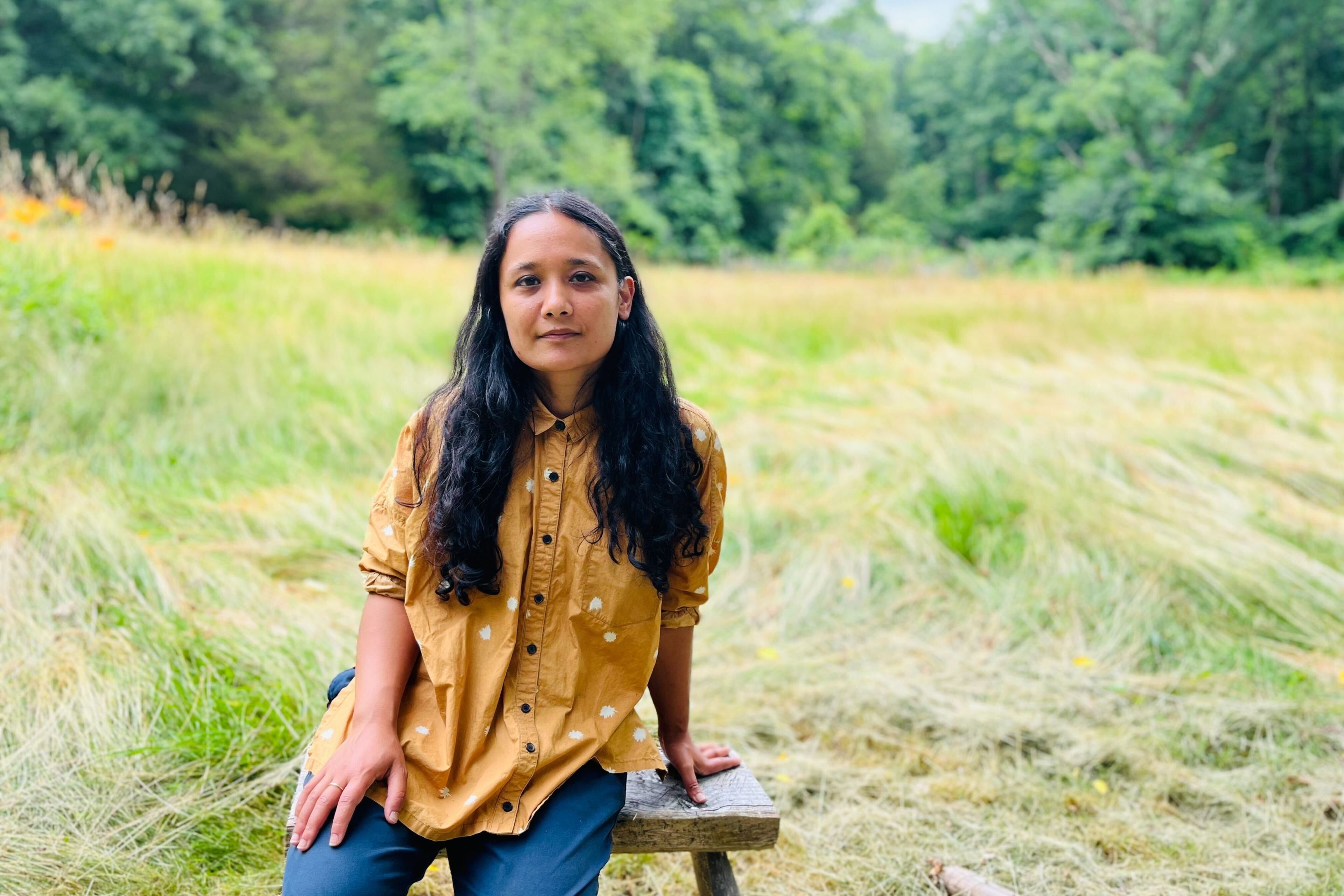 Sarahana Shrestha, the Democratic nominee for NY state Assembly District 103 in the Hudson River Valley, sits on a stool in a grassy field.