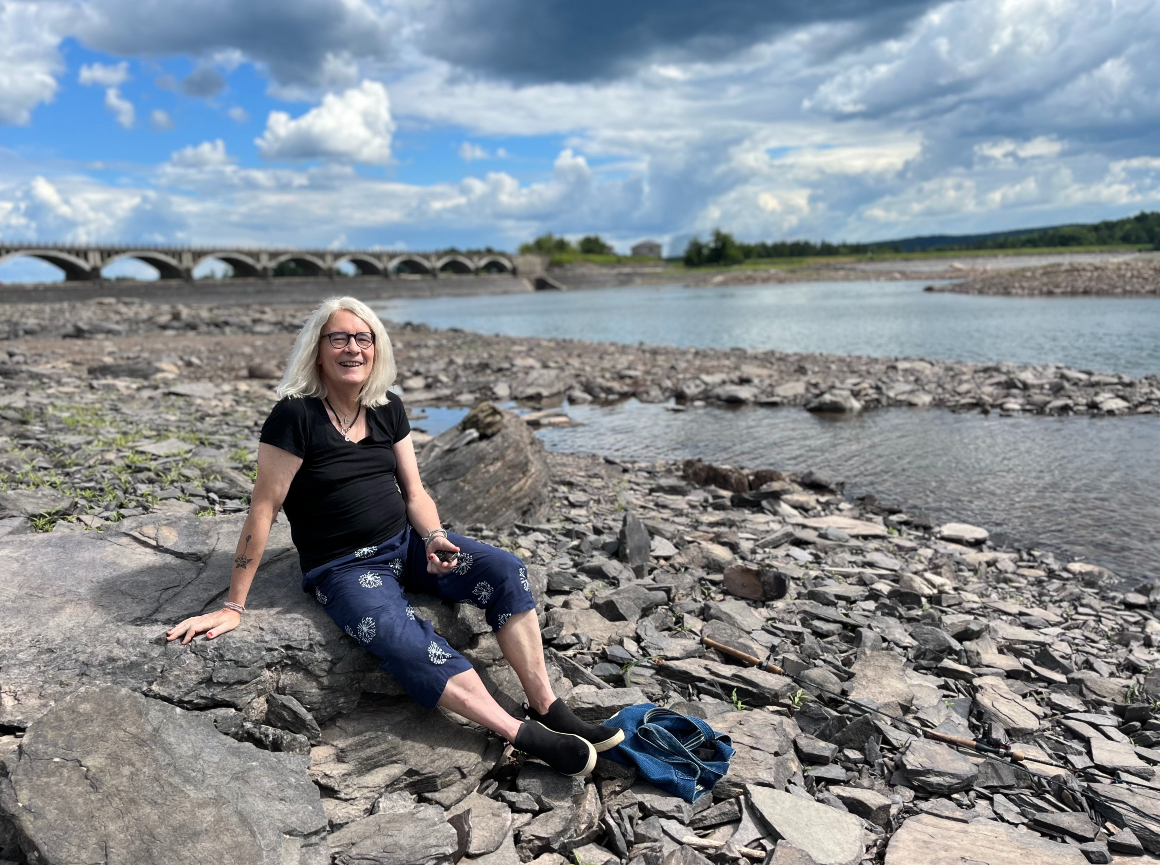 A woman in a black T-shirt and blue pants sits on the shore of the Ashokan Reservoir.