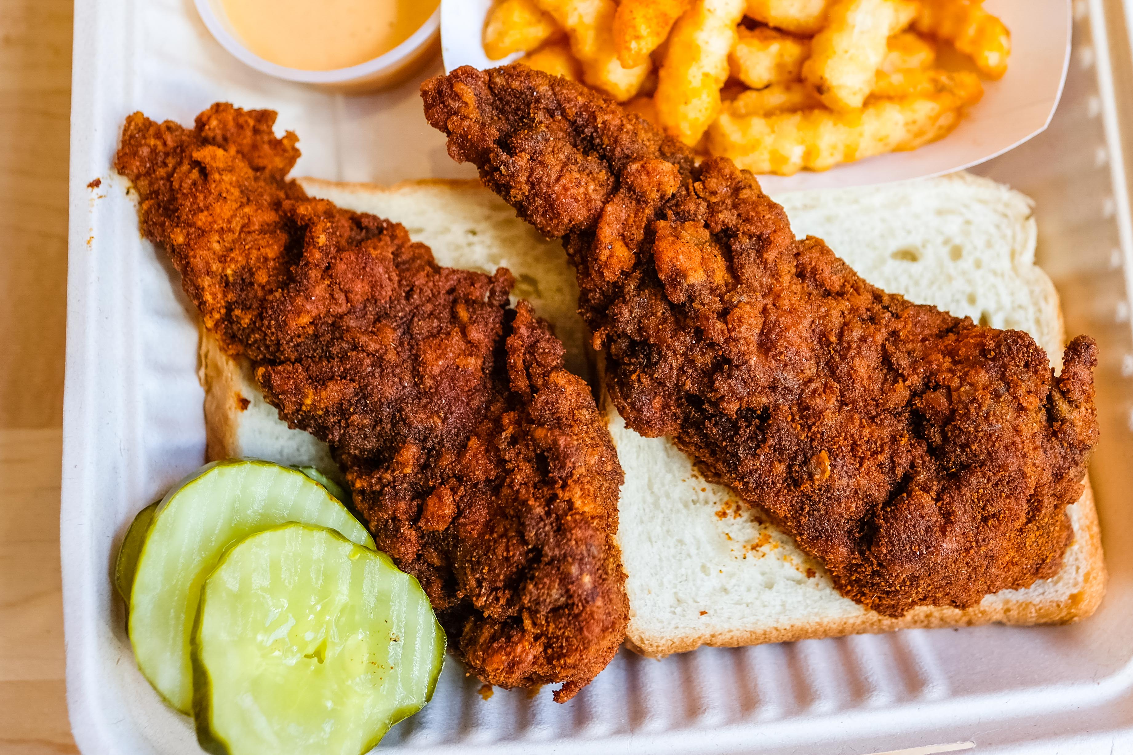 Two chicken tenders sit on a piece of white bread next to two slices of pickle and some french fries.