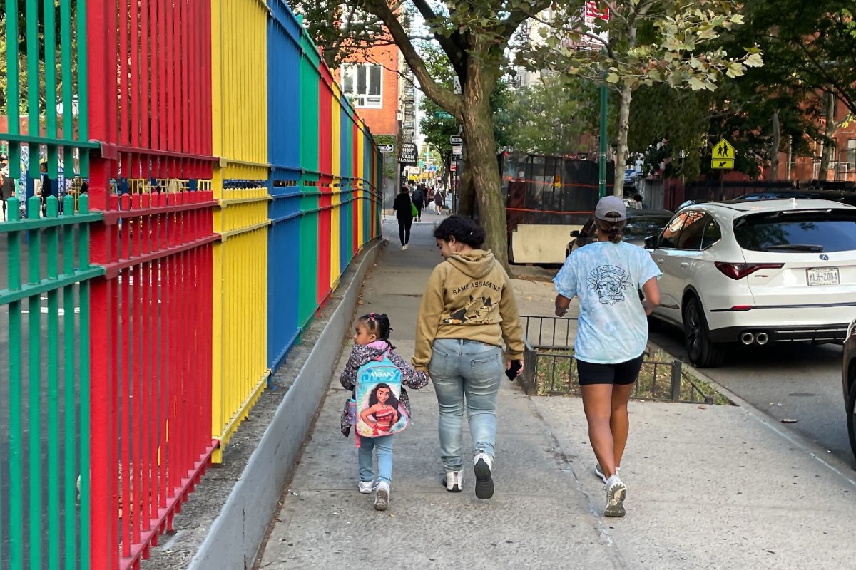 A young girl in a backpack holds the hand of an older woman as they walk on a NYC sidewalk.