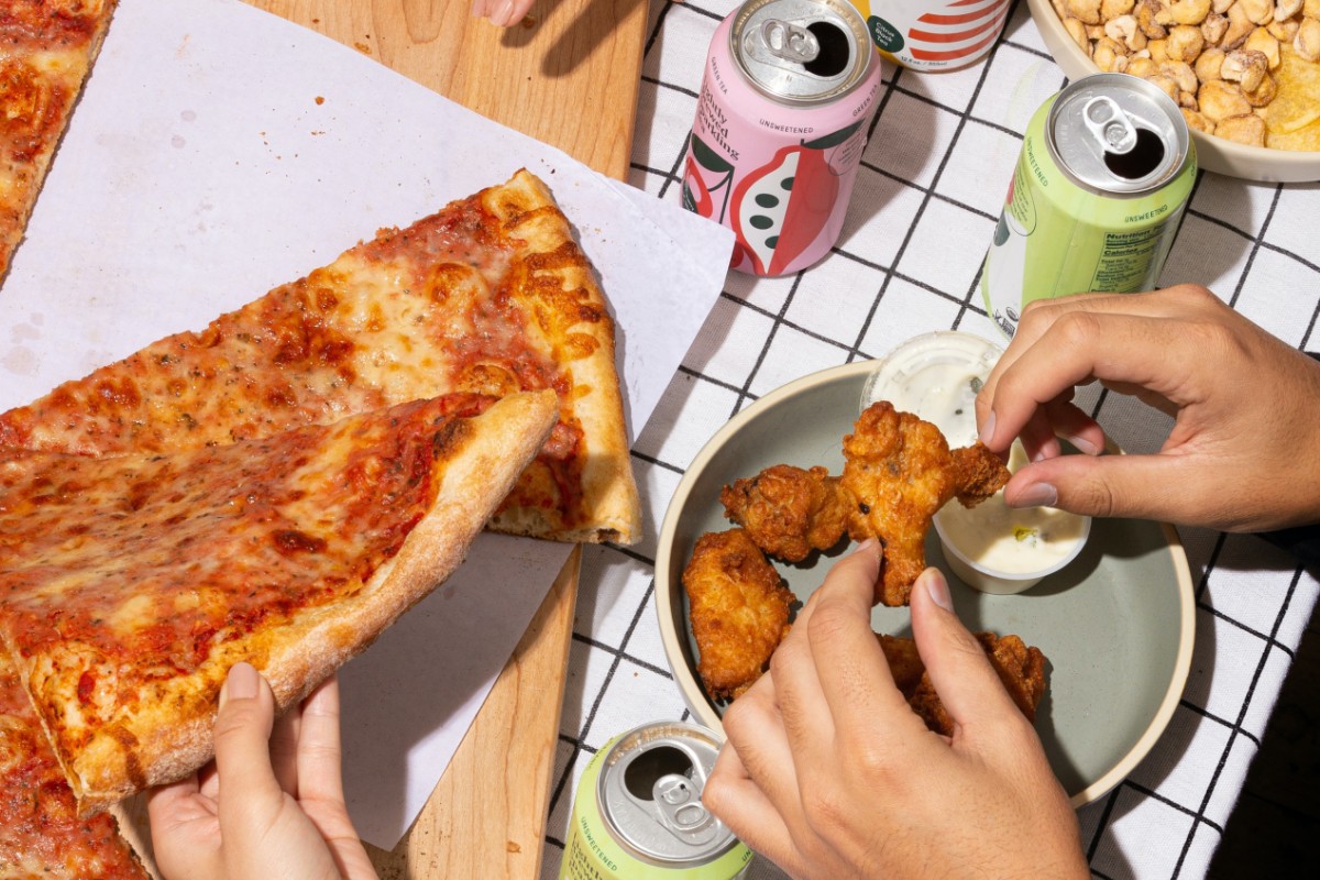 A close-up photo of two people eating pizza and chicken wings.