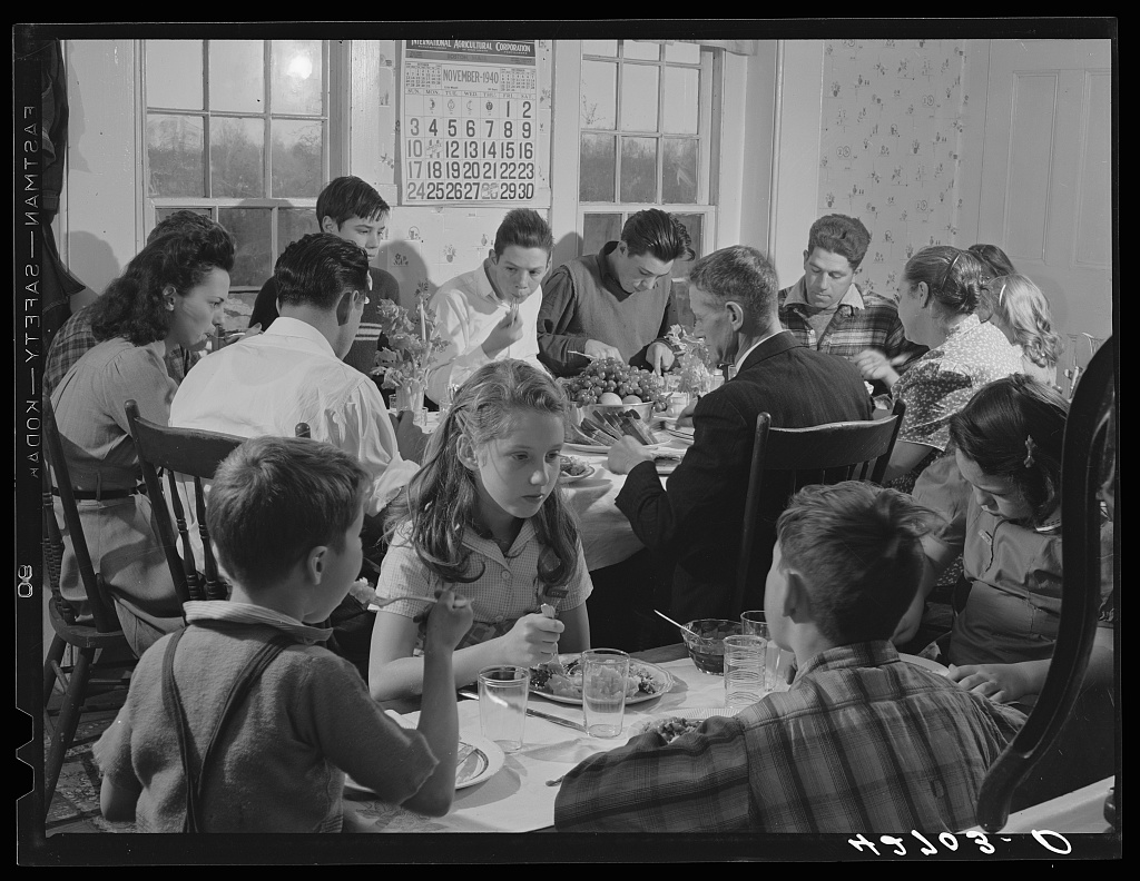 A black and white photograph of a large family siting around multiple tables eating thanksgiving dinner in 1940.