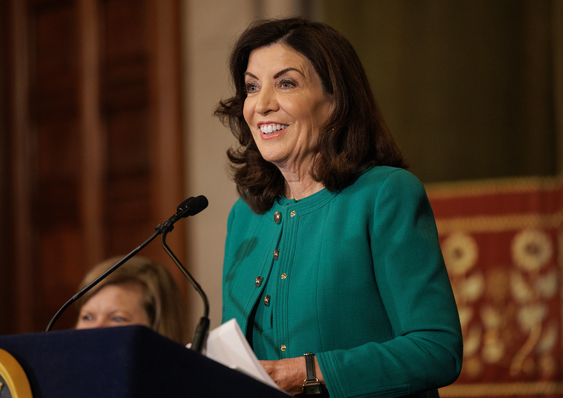 Governor Kathy Hochul smiles at a podium and sports a green jacket at a press conference.
