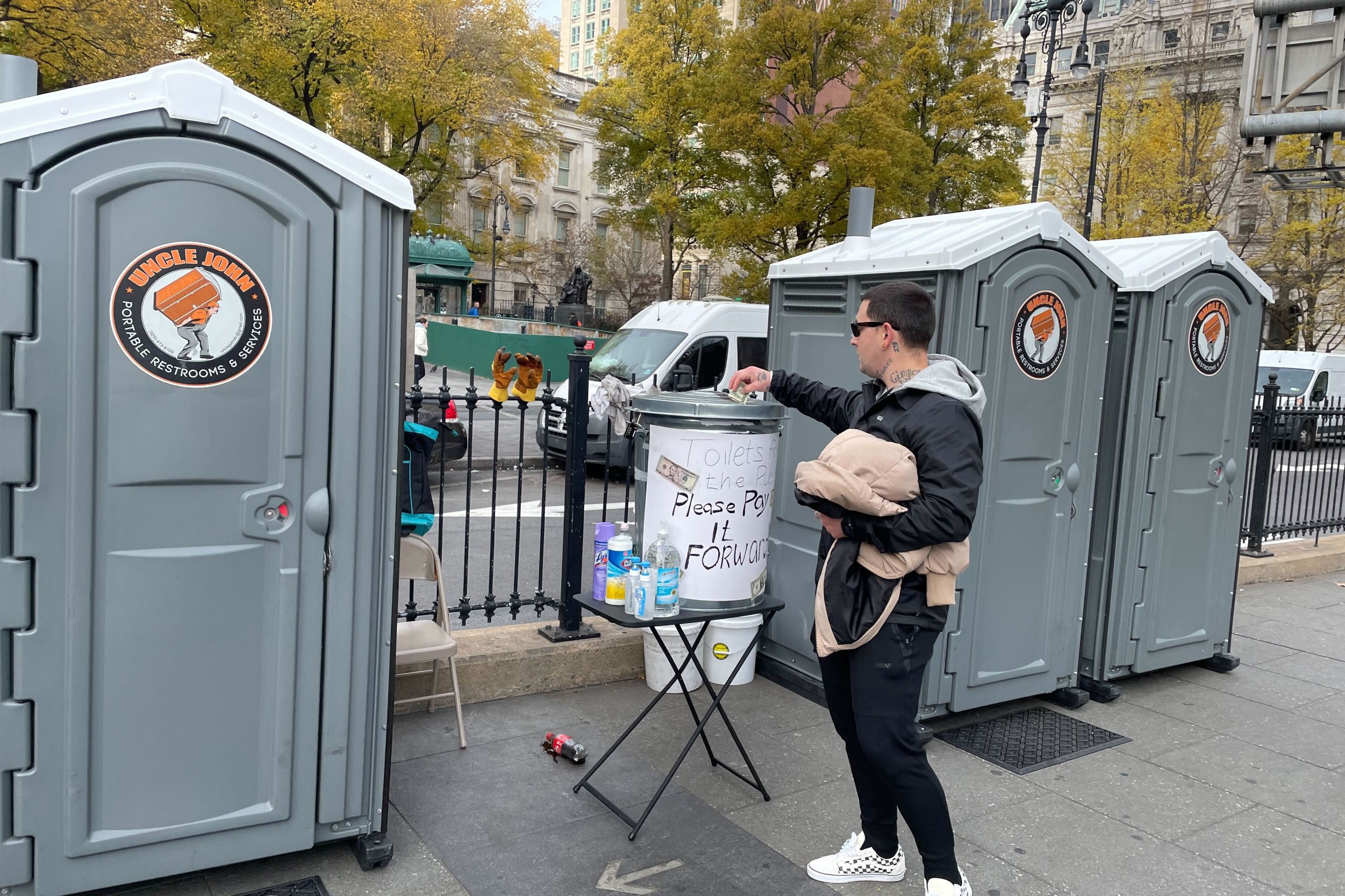 A man places a dollar bill into a trash can-turned-tip jar before using one of the port-a-potties at the base of the Brooklyn Bridge.
