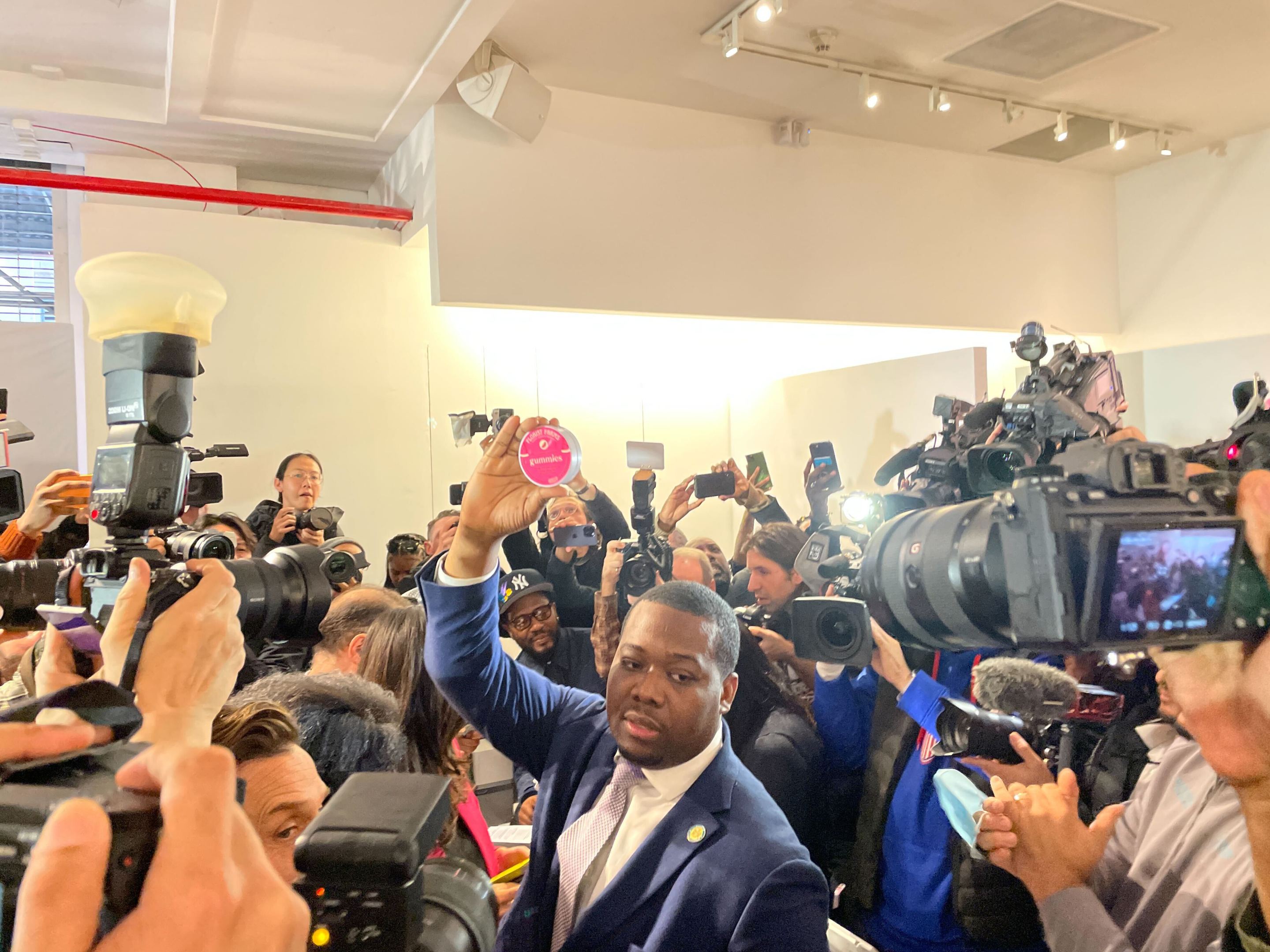 Chris Alexander wearing a suit, holding aloft a tin of legal cannabis gummies in a sea of cameras at the first legal cannabis shop at 750 Broadway. This was the first legal purchase in the state.