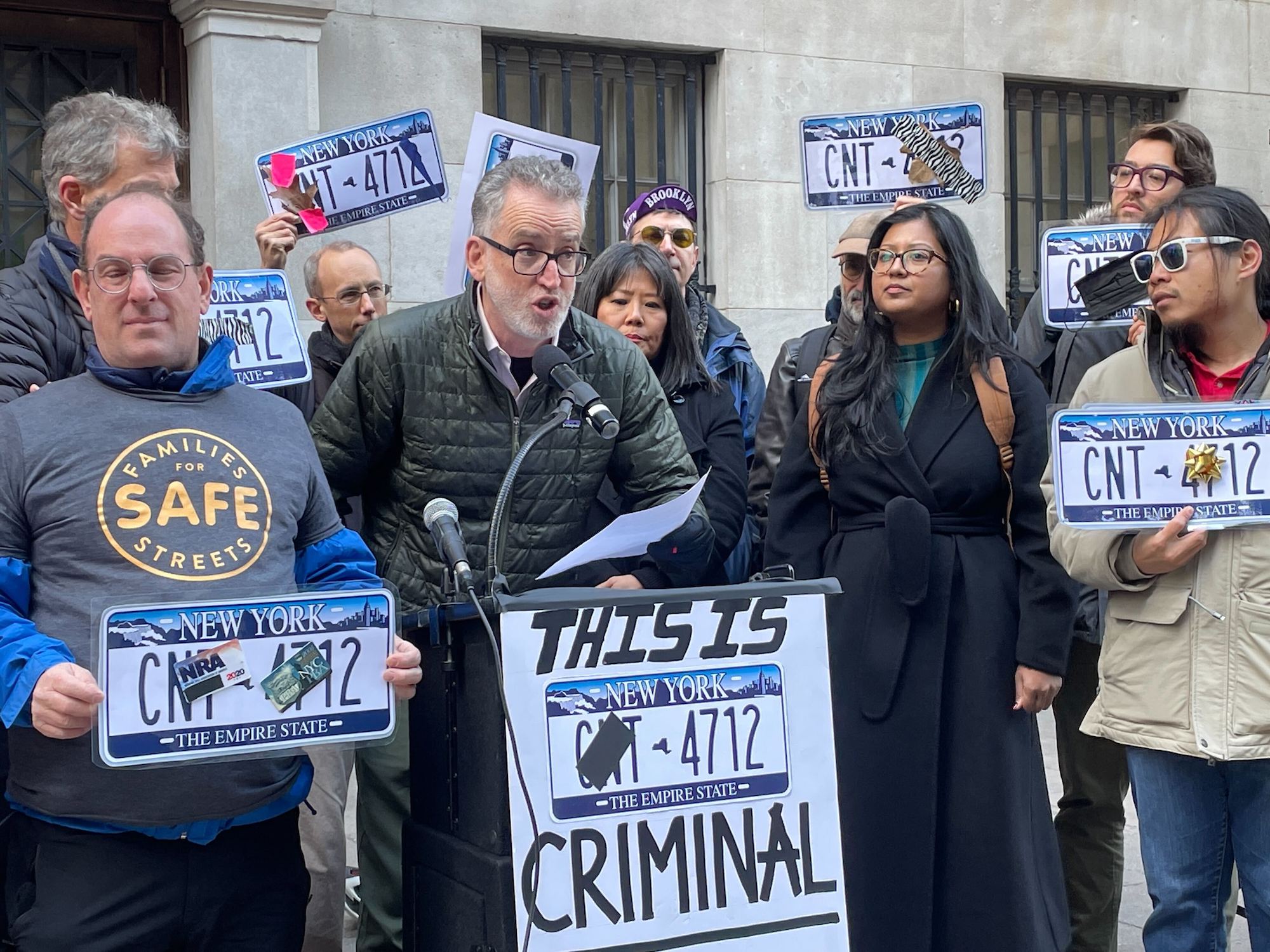 Attorney Adam White speaks into a microphone at a podium during a press conference surrounded by people holding signs of obscured license plates.