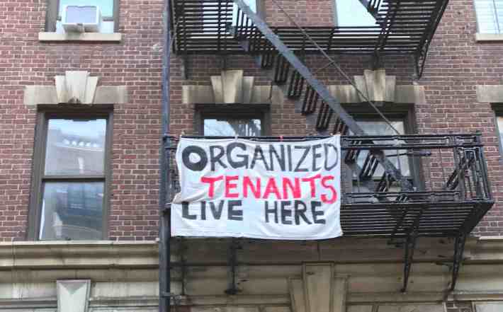 A sign hanging from the fire escape exterior of 709 West 170th Street says "Organized Tenants Live Here" in painted black and red letters.