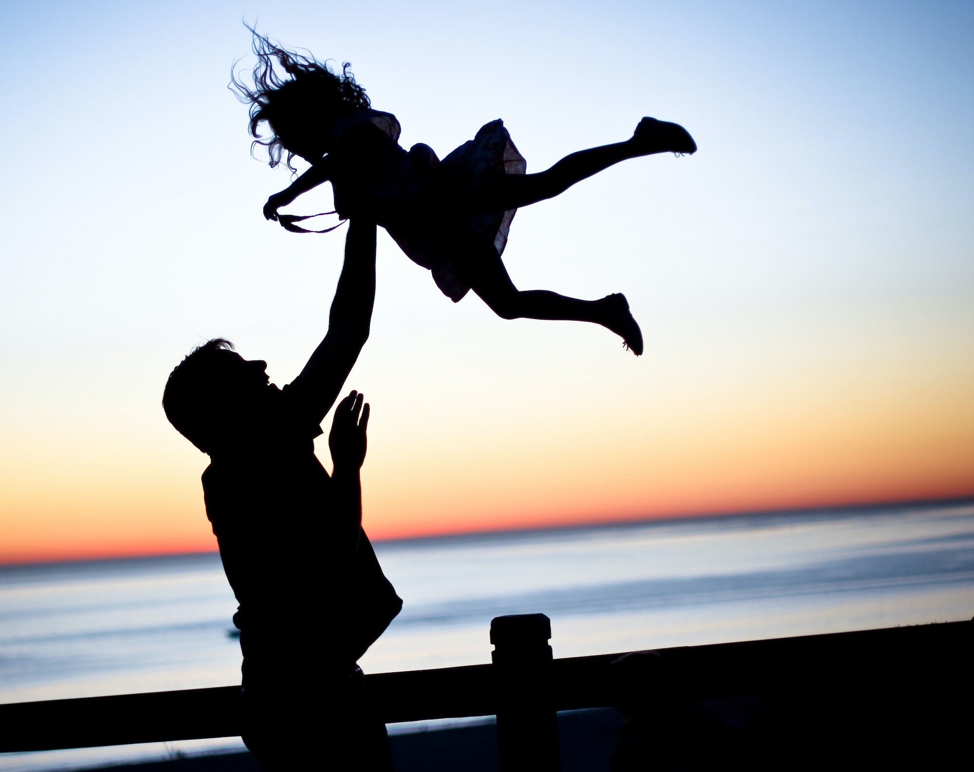 A father tosses his kid in the air playfully at sunset at the beach.