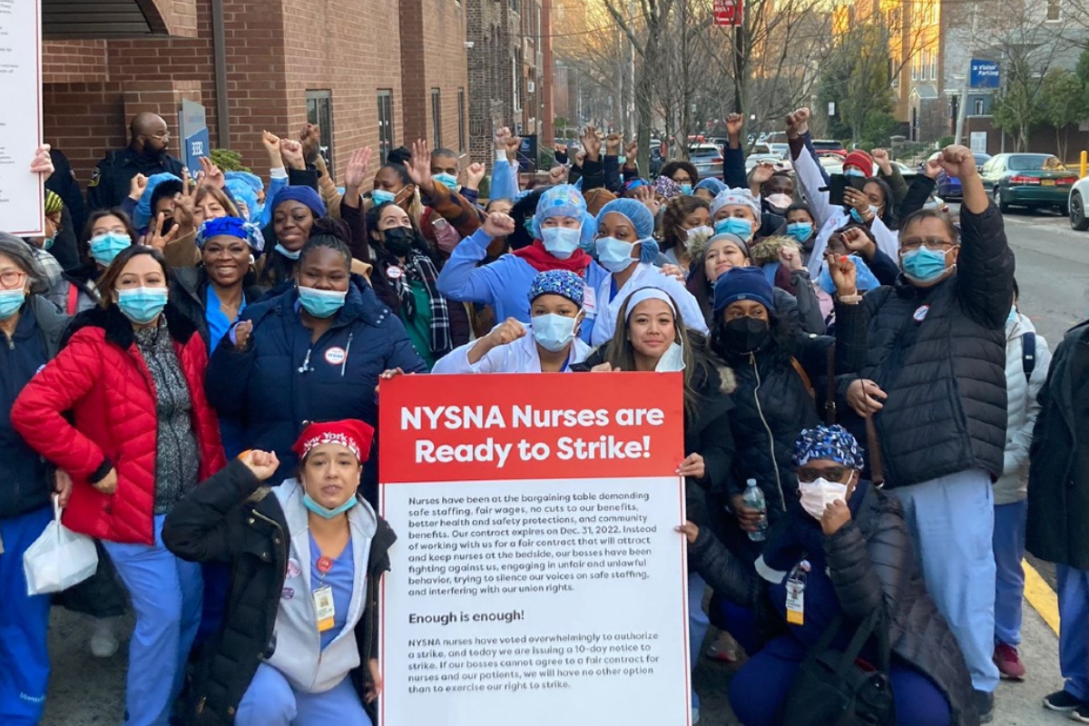 A group of more than two dozen nurses in scrubs stand together in front of a hospital, with some clutching a sign that reads, "NYSNA Nurses are Ready to Strike!"