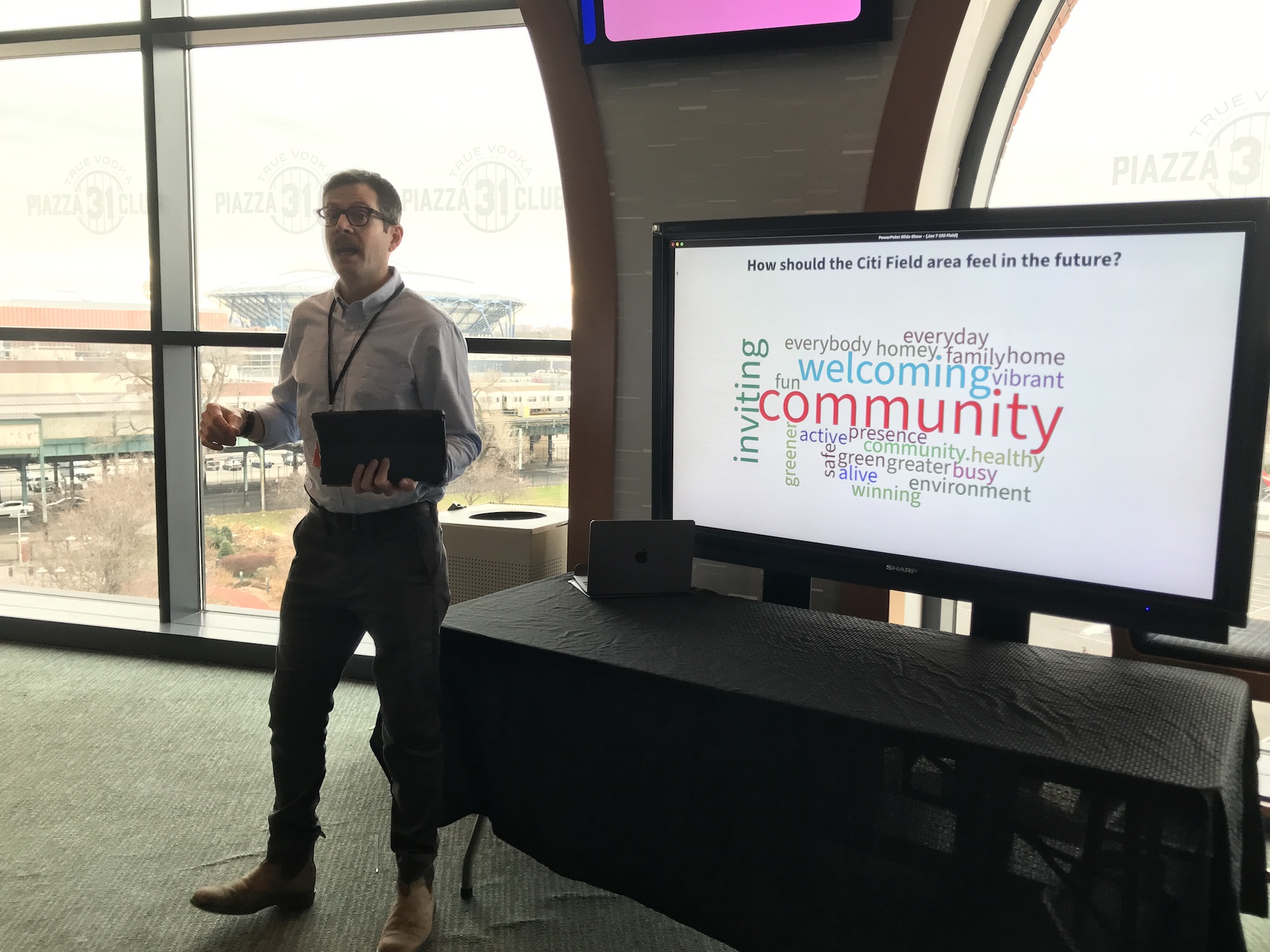 A facilitator stands in front of a flat screen TV with a word cloud, and speaks to participants at a "Visioning session" at Citi Field.