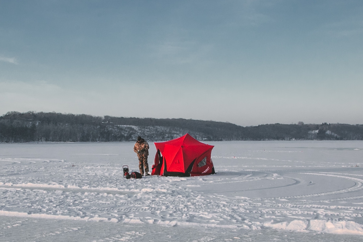An ice fisher on a frozen lake, next to a red tent.