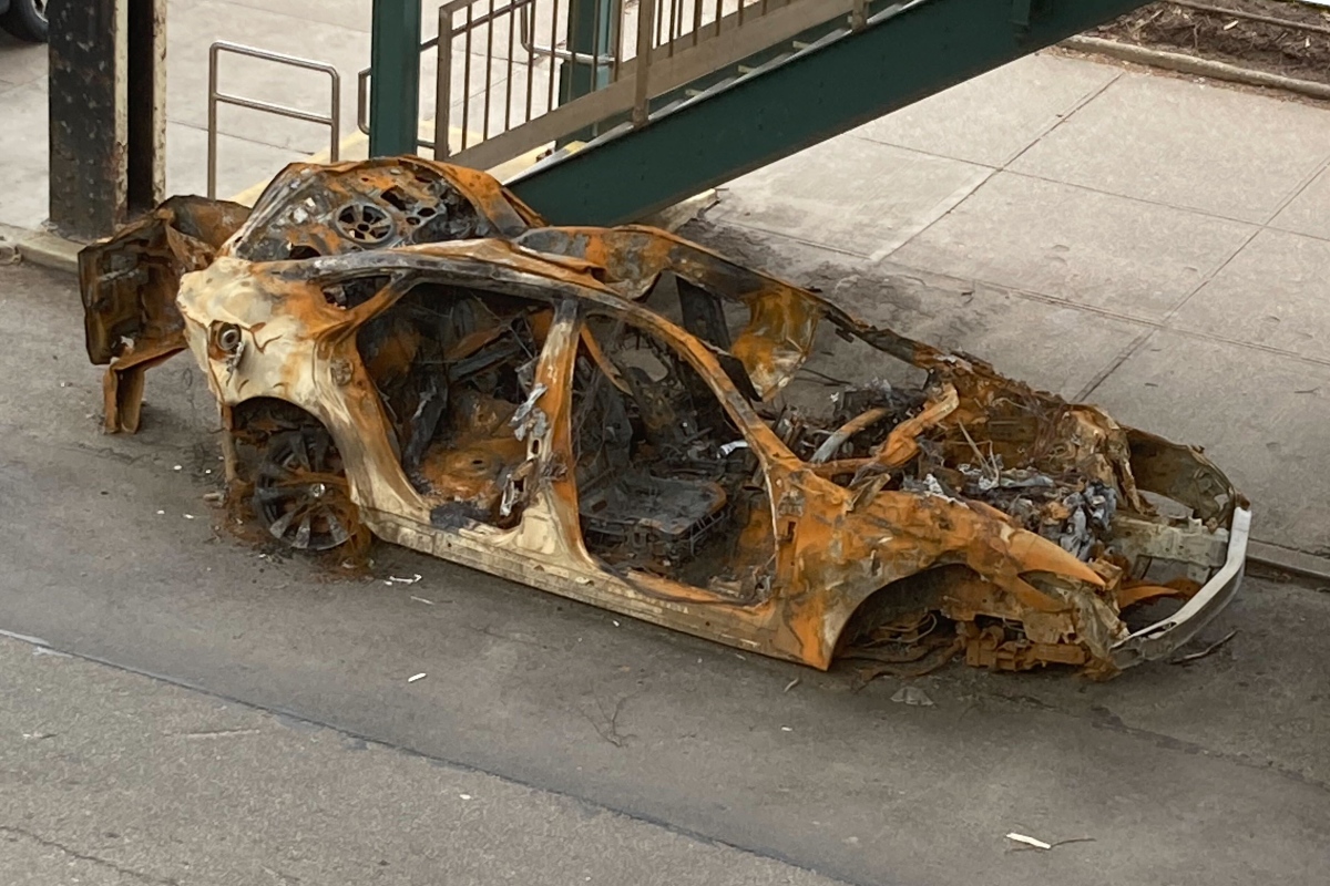 A burned out car next to the entrance of an elevated subway station.
