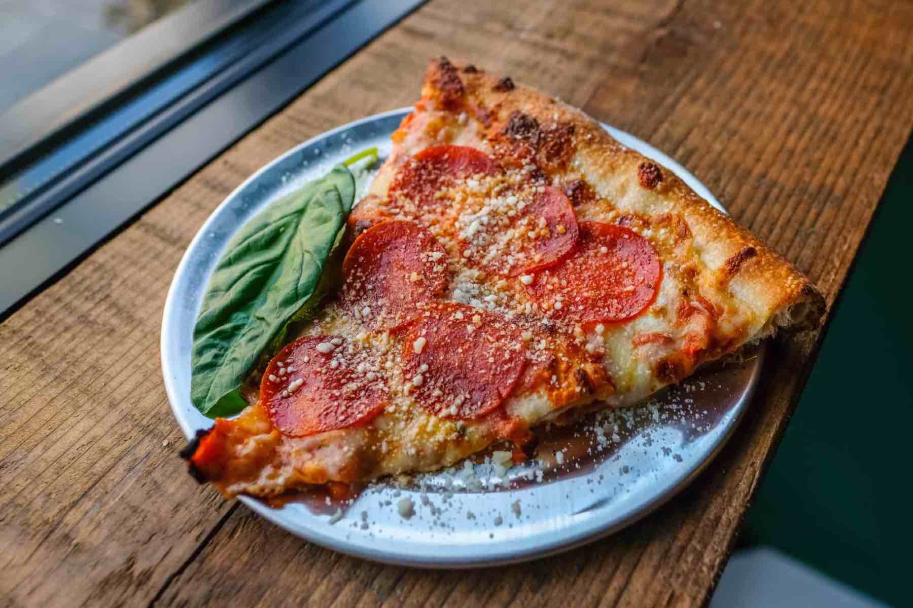 Gooey slice of pepperoni pizza on a metal plate next of a piece of basil.