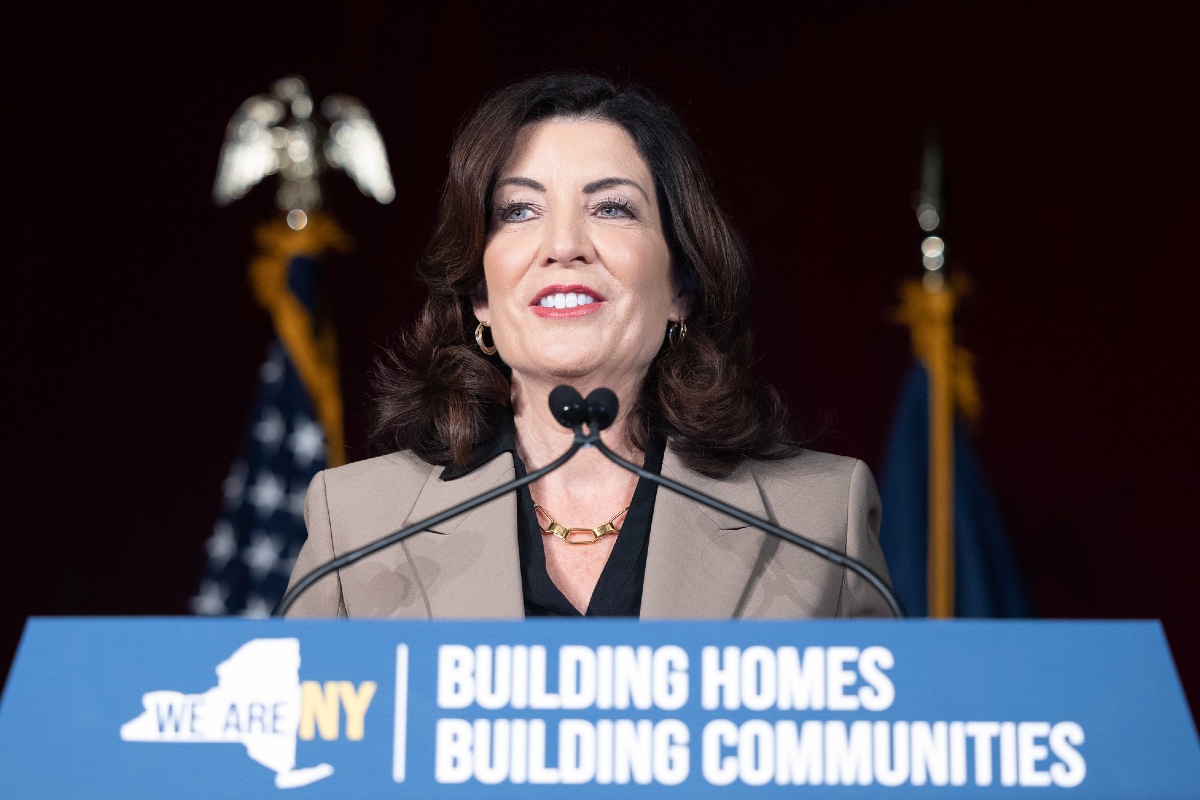 NY Governor Kathy Hochul stands behind a podium with a sign that reads, "BUILDING HOMES BUILDING COMMUNITIES."
