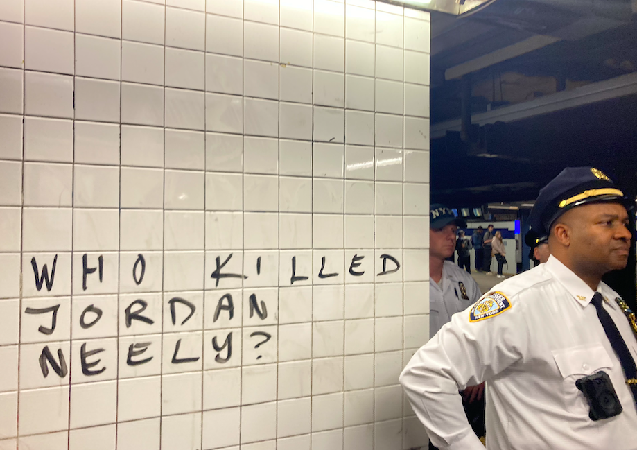A police officer stands next to subway tiles scrawled with the words, "WHO KILLED JORDAN NEELY?"