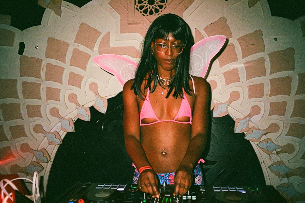 Csyan Russell, a young Black woman wearing a pink bikini top and butterfly wings, DJs at a party.