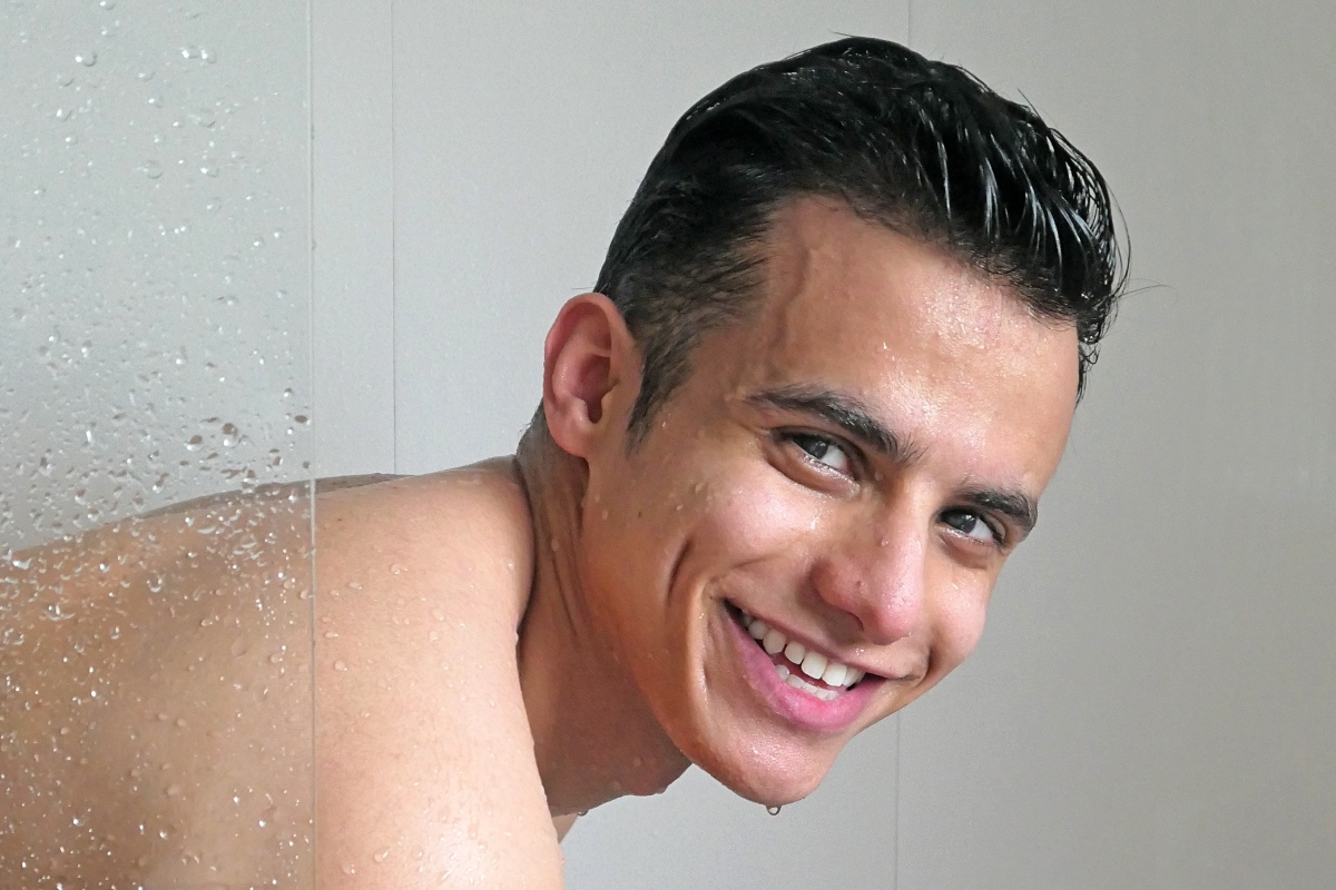 A man with slick-backed hair smiles as he peers out of a shower.