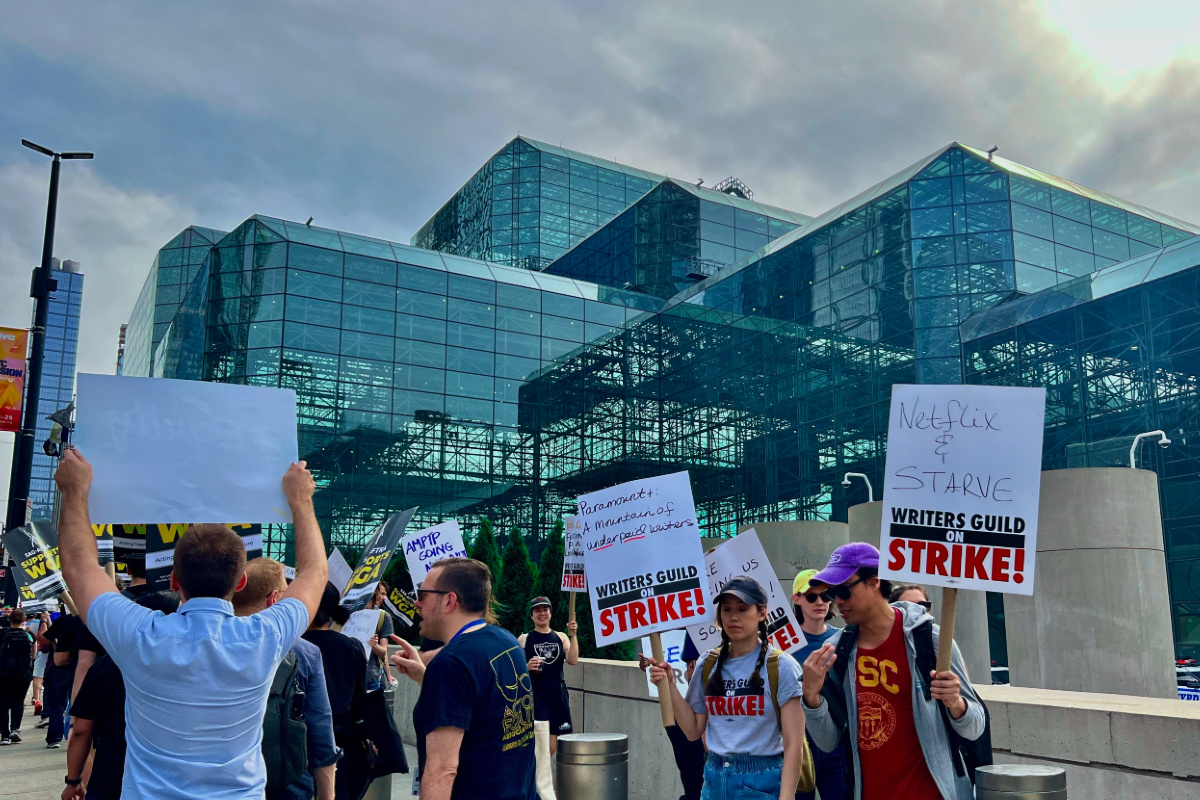A group of striking WGA writers holding signs outside of NYC's Javits Center, on a cloudy day.