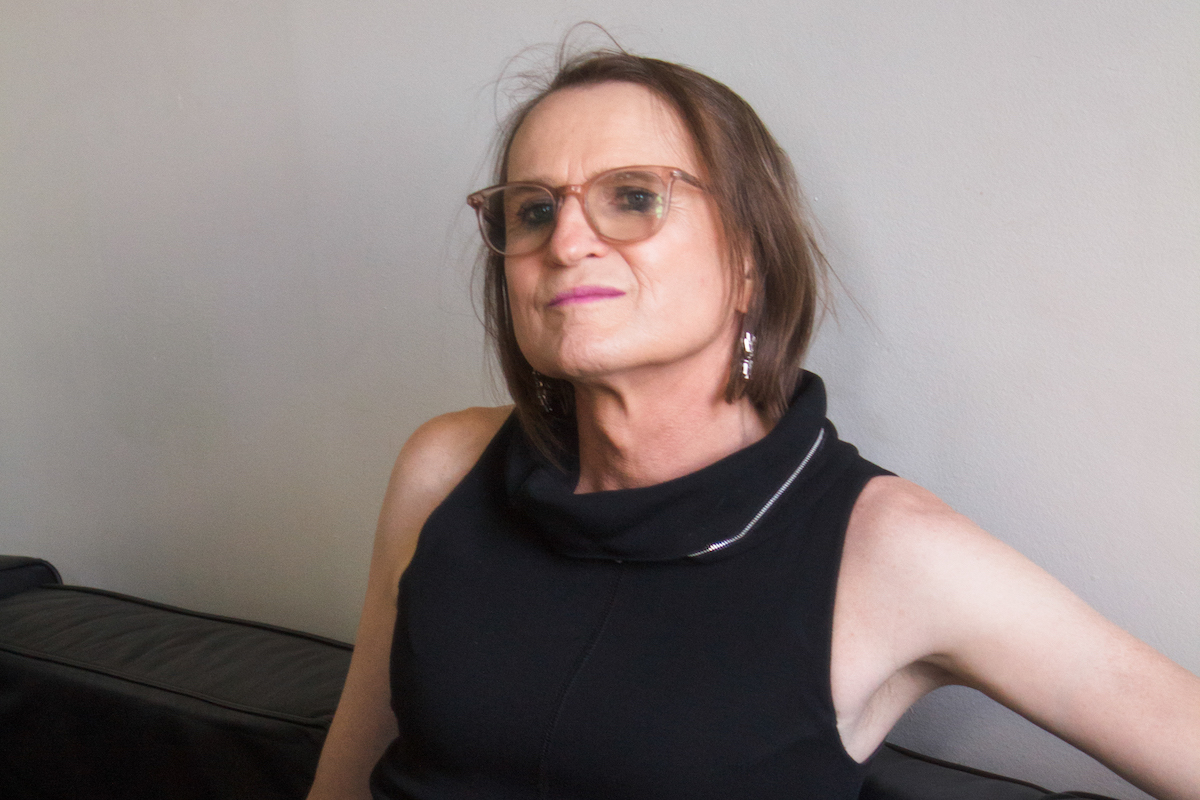 McKenzie Wark, wearing a black tank top glasses, sitting against a wall and looking off to the side.