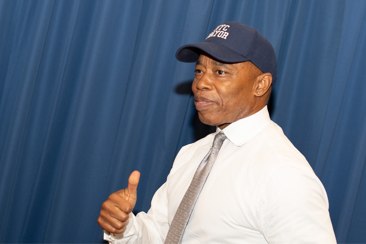 NYC Mayor Eric Adams in a blue baseball cap and white button-down, giving a thumbs up in front of a blue curtain.