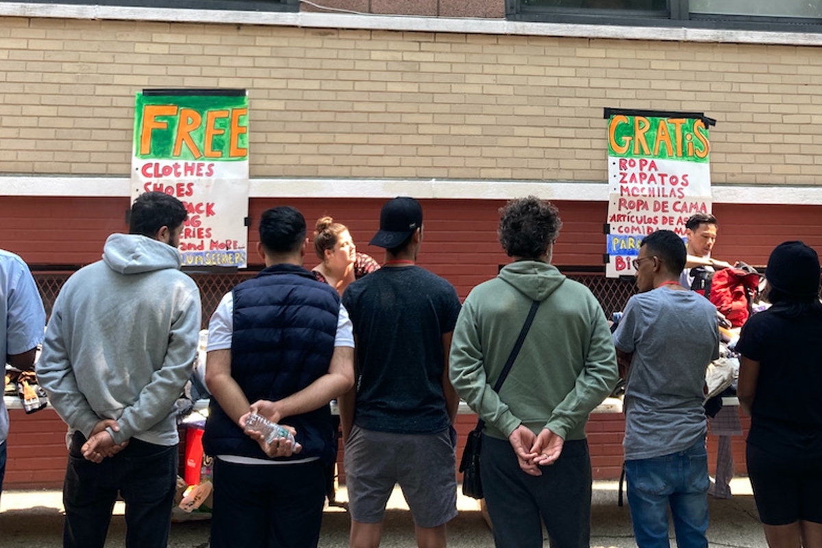 A group of people standing with their backs to the camera, in front of a clothing distribution table at a former school building.