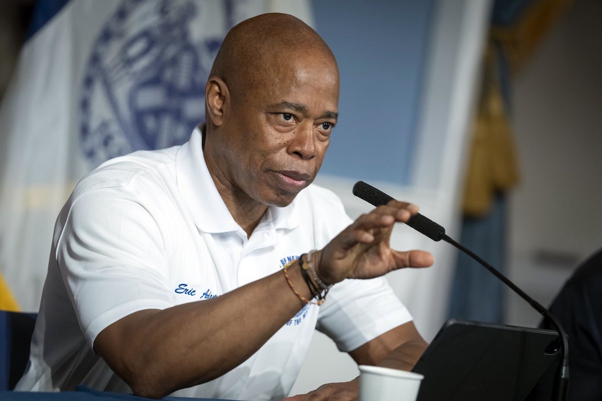 NYC Mayor Eric Adams in a white polo shirt, speaking before a podium.