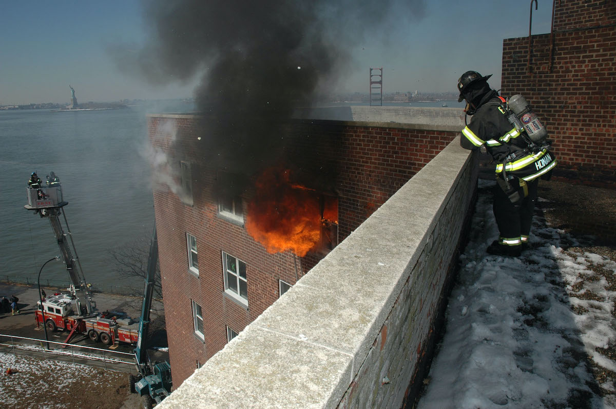 Smoke pours out of a building's window, as a firefighter watches from a nearby ledge.