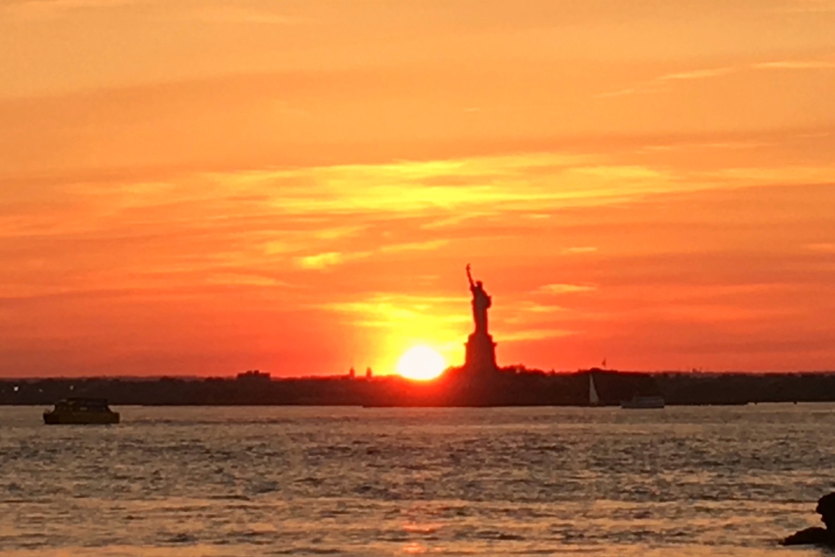 The Statue of Liberty in the distance, as the sun sets against an orange sky.