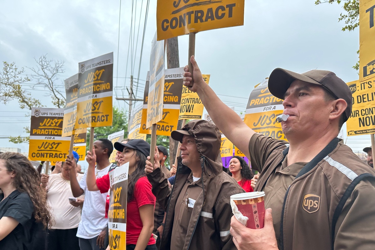 A group of people, many UPS workers in brown uniforms, hold signs saying "Just Practicing for a Just Contract"