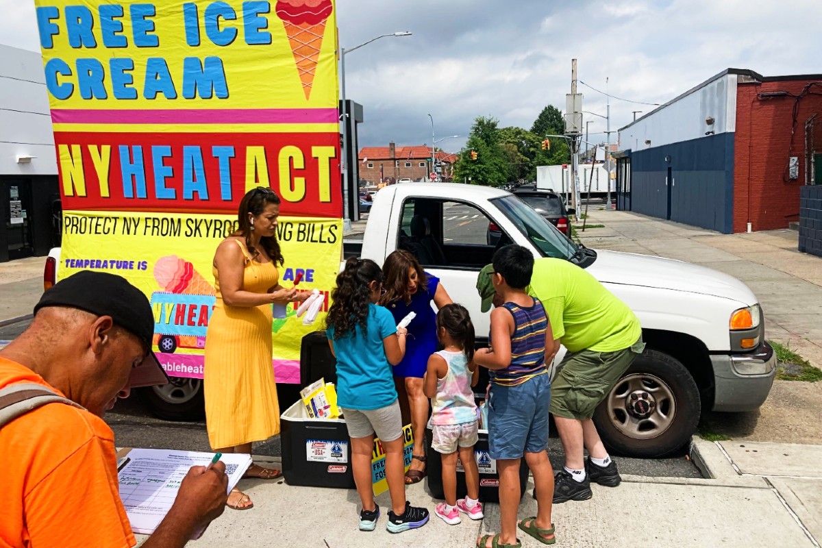A bunch of kids in front of a white truck wait for popsicles to be handed out from a woman in a yellow dress, while a man in the bottom left corner signs a petition.