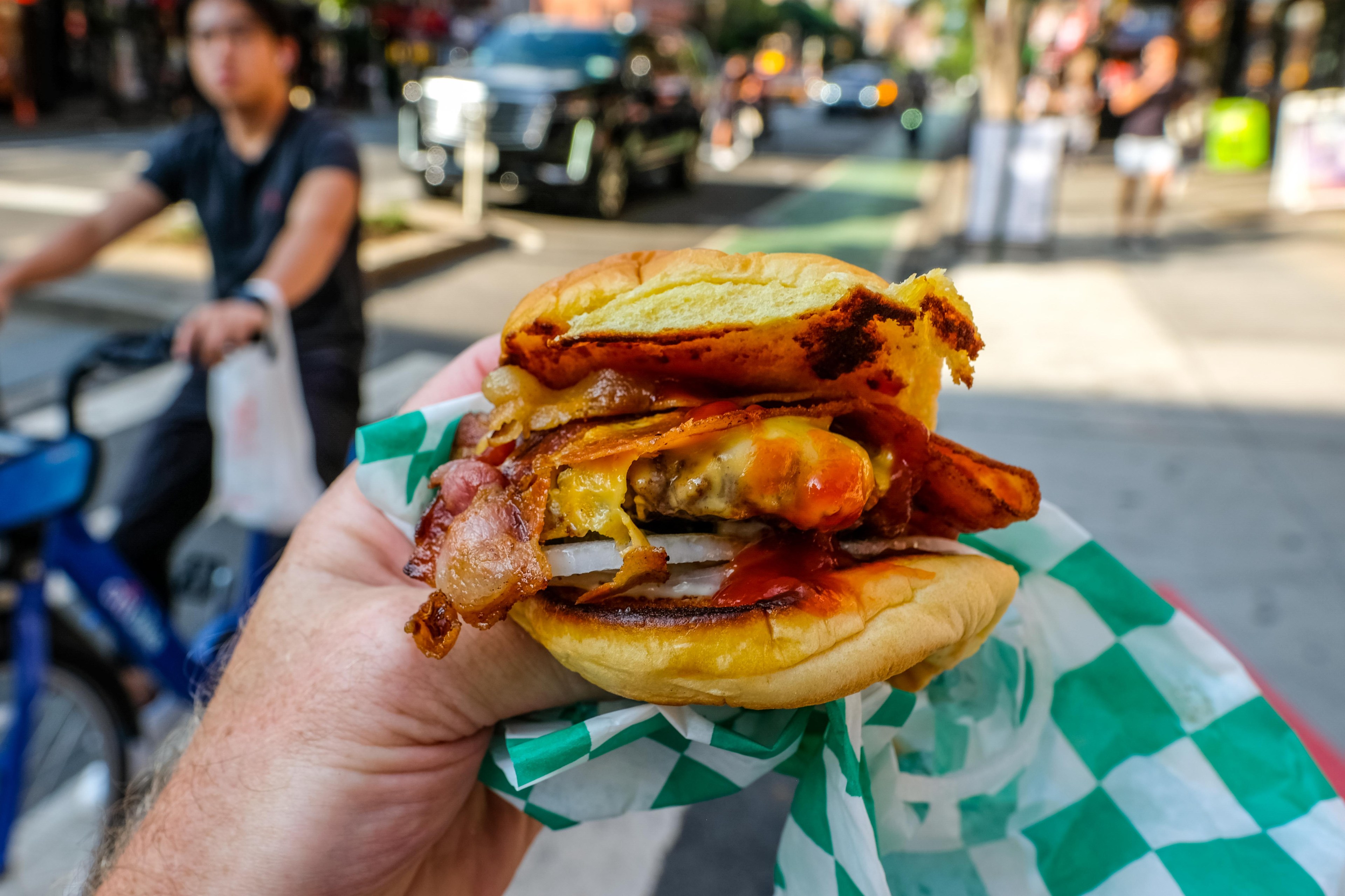 A hand holding a bacon cheeseburger on a sidewalk in New York City.